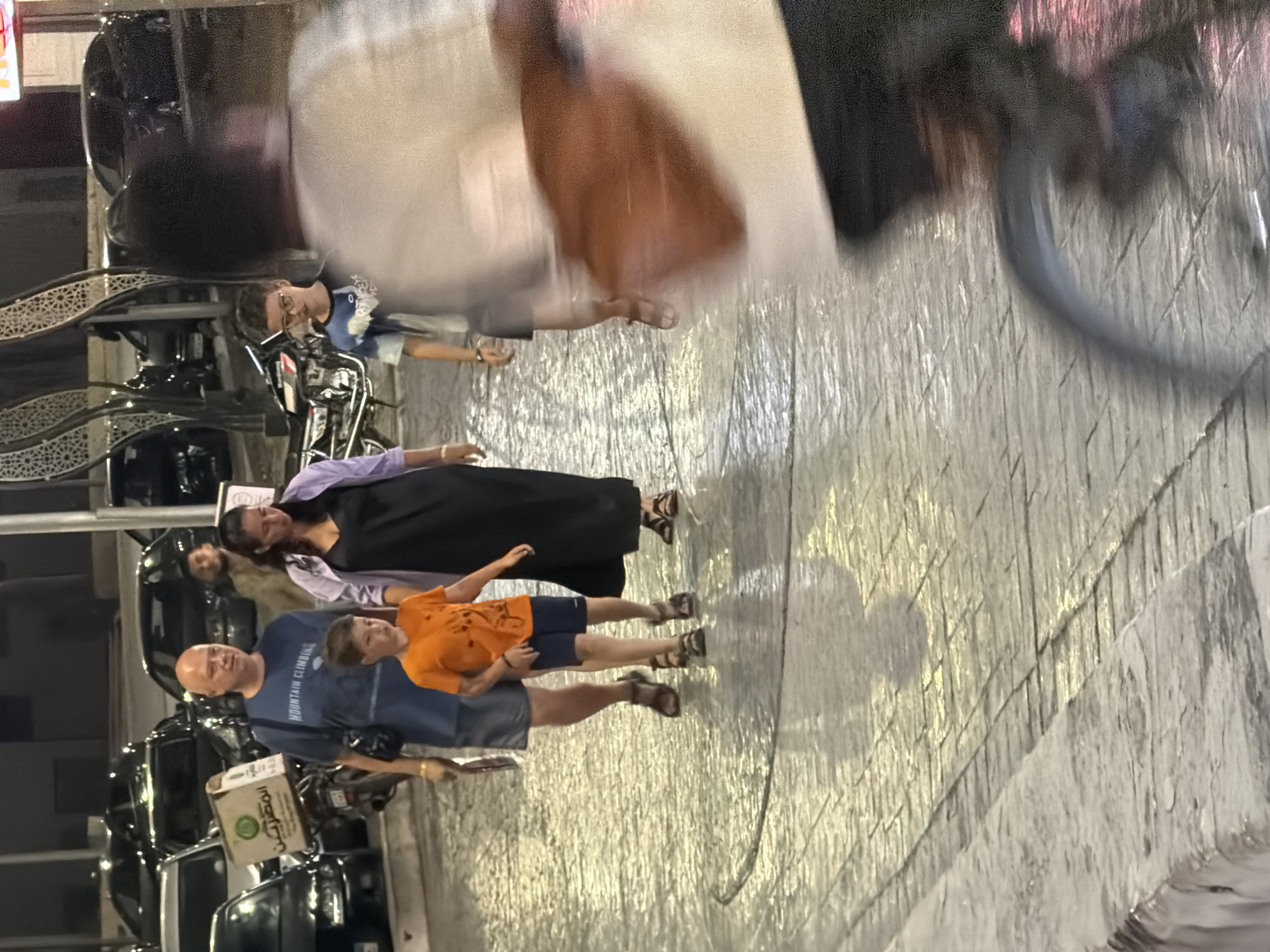 In an outdoor urban setting, likely in Sharm El Sheikh, Egypt, a group of four individuals is walking on a cobblestone pavement. The group consists of a bald man in a blue t-shirt, a woman in a black dress and a light purple jacket, a young boy in an orange t-shirt and dark shorts, and another boy in a t-shirt with a blue and white pattern and glasses. They appear to be walking in the same direction, with the two boys in the middle of the group. The man is carrying a box on a motorcycle parked to his left, with Arabic writing visible on the box. The woman is walking next to the man, and the two boys are in front of them. In the background, several cars are parked, and some streetlights are visible, casting reflections on the wet pavement. The lighting suggests it is evening or nighttime. A person on a bicycle is partially visible in the foreground, moving out of focus.