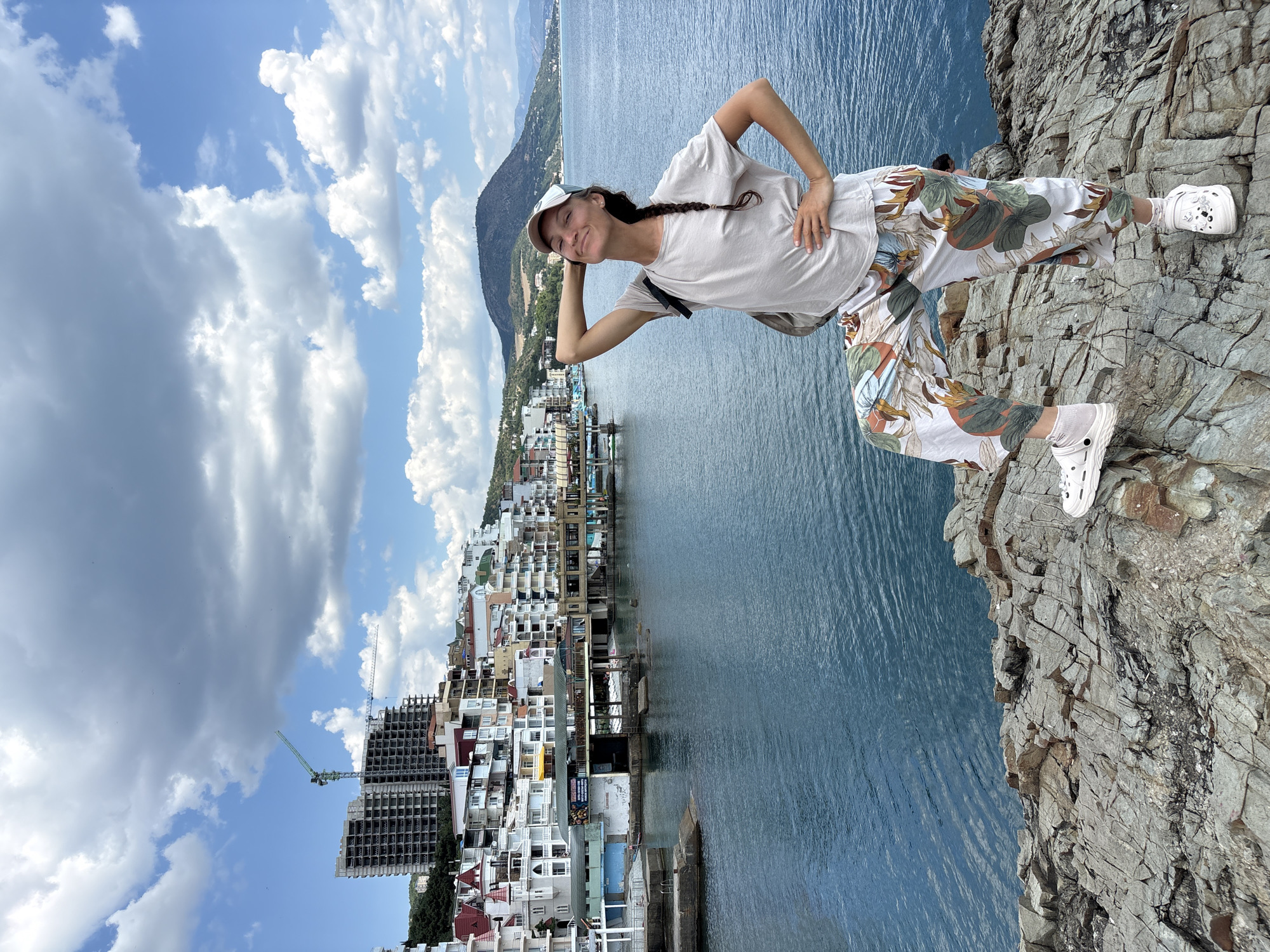 A woman with braided brown hair, wearing a light-colored t-shirt, patterned pants, a white baseball cap, and white Crocs, poses on a rocky outcrop overlooking the Black Sea in Kiparisnoe, Ukraine. She has one hand behind her head and the other on her hip, with her right leg propped up. The sea is a deep blue, and in the background, a town with white buildings stretches along the coastline, with a construction site featuring a crane visible on a hillside. A large, dark green mountain rises in the distance. The sky is filled with dramatic white and gray clouds.
