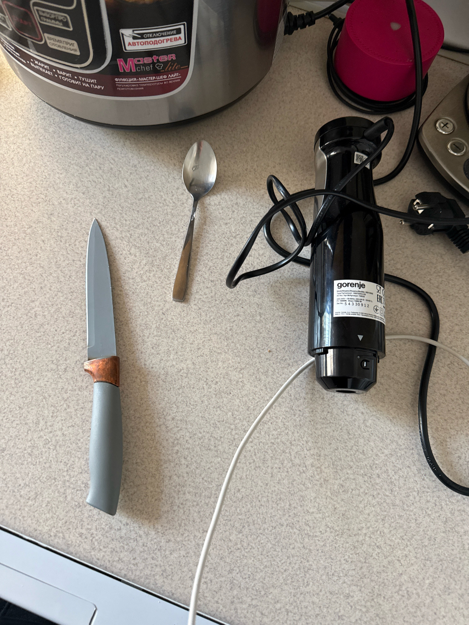 The image displays a kitchen counter with several items on it. In the foreground, a grey-handled knife with a copper bolster and a silver blade is positioned vertically on the left. To its right, a single metal spoon is laid horizontally. Further to the right is a black immersion blender with a white cord. The blender has a label with "gorenje" prominently displayed, along with product information. In the background, a large silver appliance, possibly a multi-cooker, is visible, with various labels in Cyrillic text. A red cylindrical object and electrical cords are also present. The scene appears to be an indoor domestic setting. There is no visible indication of location within Chișinău, Moldova, or any specific actions or interactions taking place beyond the arrangement of the objects.