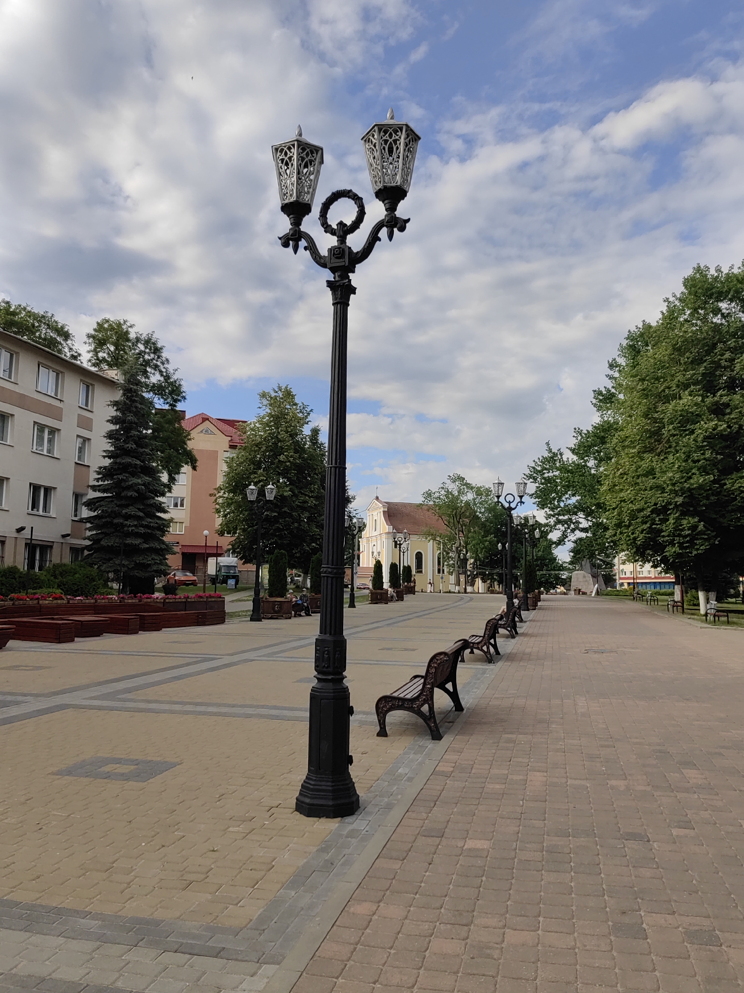 This media file depicts a serene outdoor scene, likely a town square or park, under a partly cloudy sky. The time of day appears to be late afternoon or early evening, given the soft lighting and long shadows. The weather is pleasant, with a gentle breeze suggested by the clouds.
The main subjects are the elements of the urban landscape. A prominent, ornate black lamppost stands in the foreground, featuring two decorative lamps with intricate scrollwork. To its left, a modern building with cream-colored walls and multiple windows is visible, along with a tall, dark green conifer tree. Further down the paved walkway, a row of dark brown park benches with ornate metal legs offers seating.
The setting is a pedestrian walkway paved with tan and grey interlocking bricks, bordered by darker red bricks on the right. Along the left side of the walkway, there are raised wooden planters filled with red flowers and greenery. Beyond the planters, more buildings are visible, including a building with a red-tiled roof and a light yellow building with arched windows, possibly a church or public building. Green trees line the walkway on both sides, creating a sense of natural beauty within the urban environment.
There are a few people visible, casually sitting on some of the benches in the distance, suggesting a peaceful atmosphere and a place for relaxation. No specific activity or event is clearly taking place, implying a typical day in this public space.
Notable details include the architectural style of the lamppost and the church-like building, which might hint at a European location. The overall mood is tranquil and inviting, with the well-maintained pathways, benches, and greenery creating an appealing public space. There is no visible text in the image.