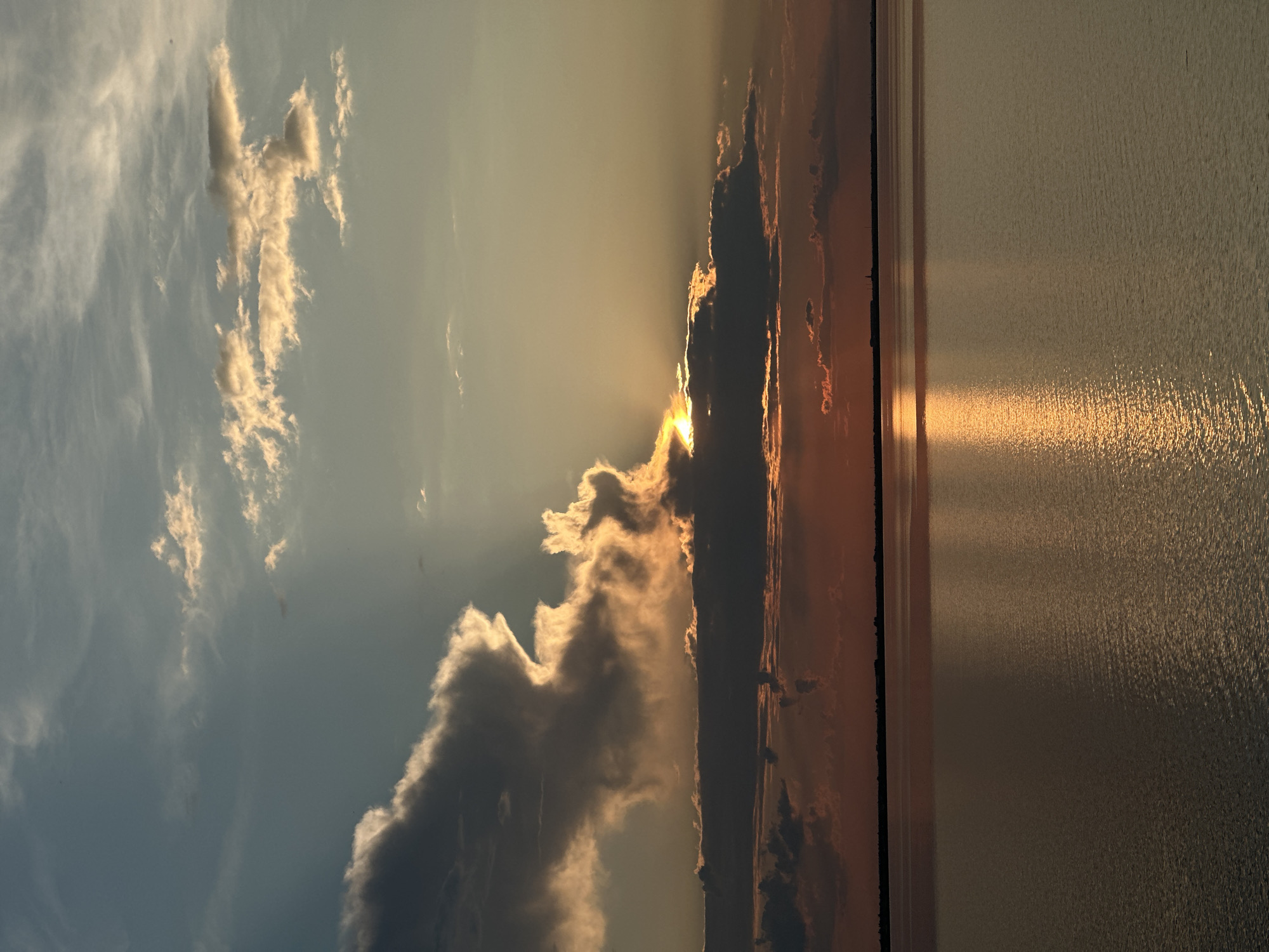 The image depicts a serene sunset over a large body of water, likely Lake Kokshetau, given the specified location. The sky is a gradient of blues, grays, and oranges, with dramatic clouds partially obscuring the setting sun. Rays of sunlight pierce through the clouds, casting a golden glow on the water's surface. The water itself is calm, reflecting the vibrant colors of the sky. The horizon is marked by a dark, silhouetted line of land, possibly indicating the shore with distant structures or trees. There are no people or animals visible in the frame. The overall atmosphere is tranquil and picturesque.