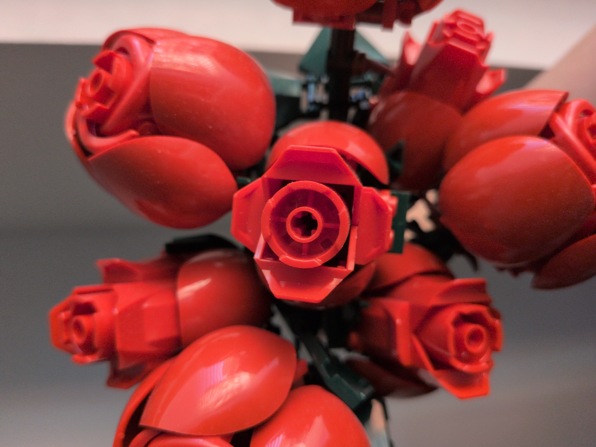 The image displays a detailed, close-up, high-angle view of a cluster of red roses, meticulously constructed from Lego bricks, situated in the city of Unknown, Unknown. Multiple fully bloomed and partially opened rose constructions are visible. The central rose is sharply in focus, revealing its internal structure, including a visible central cross-shaped axle hole. Other roses feature distinct, layered red petals with glossy surfaces reflecting ambient light. Subtle green Lego elements, likely representing stems or leaves, are faintly discernible between the red rose structures. The background is a uniformly blurred gradient from dark gray to off-white, indicative of an indoor setting. No human figures or active interactions are depicted within the scene.