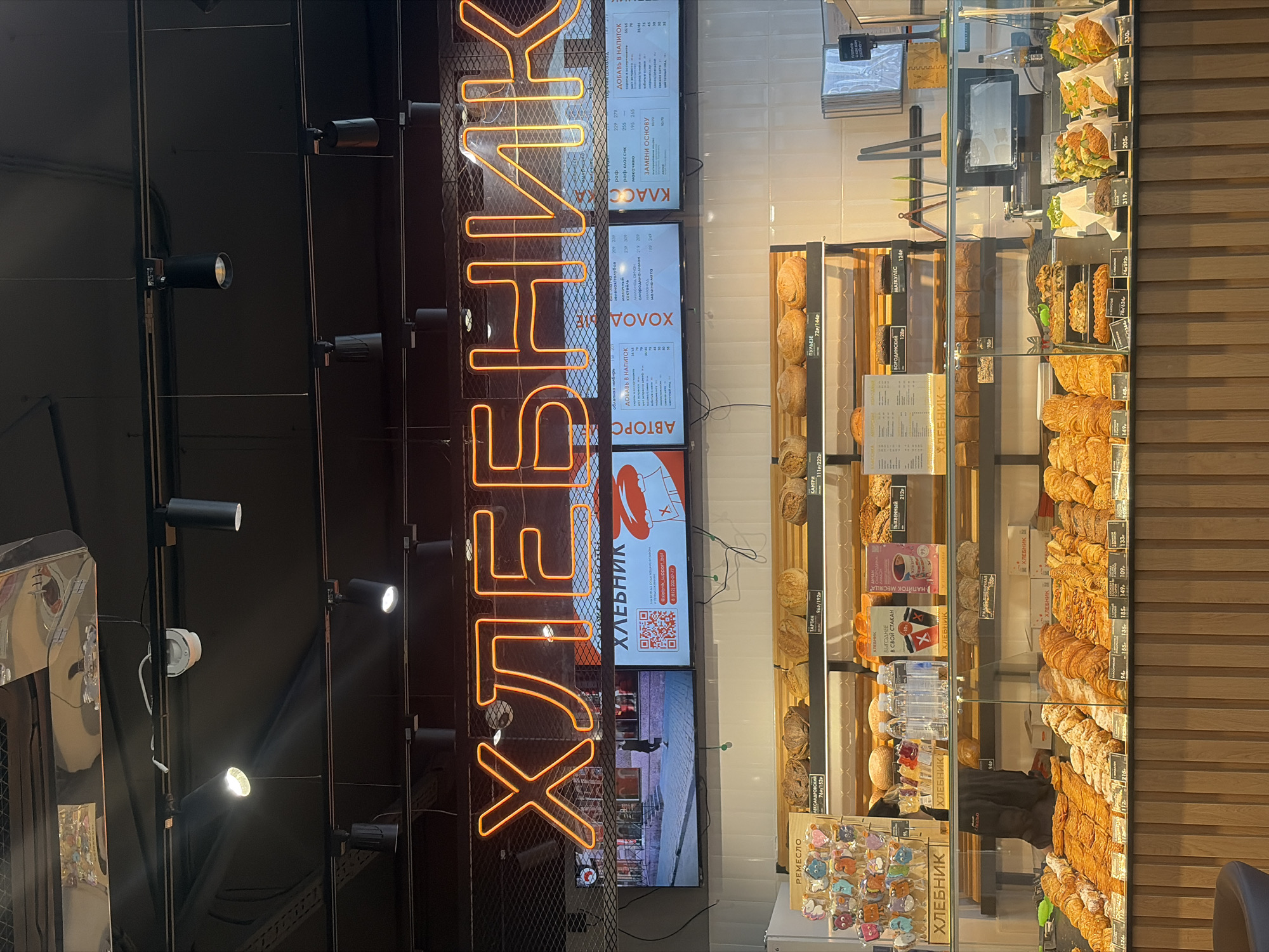 This image captures the interior of a "ХЛЕБНИК" (Khlebnik) bakery located in Saint Petersburg, Russia, identifiable by the prominent "812" telephone area code displayed on a digital menu board. The scene is dominated by a modern commercial bakery counter under a large, orange neon sign spelling "ХЛЕБНИК" against a dark ceiling.
Below the neon sign, three digital screens display menu items, pricing in Russian rubles (e.g., "152p", "192p"), and store information, including a QR code and a Saint Petersburg contact number. The upper section of the image also shows a dark ceiling with exposed black track lighting fixtures and a white security camera.
The central feature is a multi-tiered glass display counter. The upper shelves, positioned behind the glass, hold various loaves of rustic bread, labeled with terms like "РЕМЕСЛО" (Remeslo), "ТАРТИН" (Tartin), "ПУЛЕЗЕ" (Puleze), "БОРОДИНСКИЙ" (Borodinsky), and "МАТУШКА" (Matushka). The lower, brightly illuminated glass shelves showcase an extensive selection of pastries, including rows of croissants, various sweet buns, and individually wrapped savory items such as sandwiches and salads, each with small price tags.
Behind the counter, additional bread is stacked on shelves, and a section of white tiled wall is visible. A black point-of-sale monitor and various serving utensils are present on the right side of the counter. The front of the display counter features a base constructed from vertical light-colored wooden slats. A reflection of a person in a dark jacket is discernible in the glass of the display case.