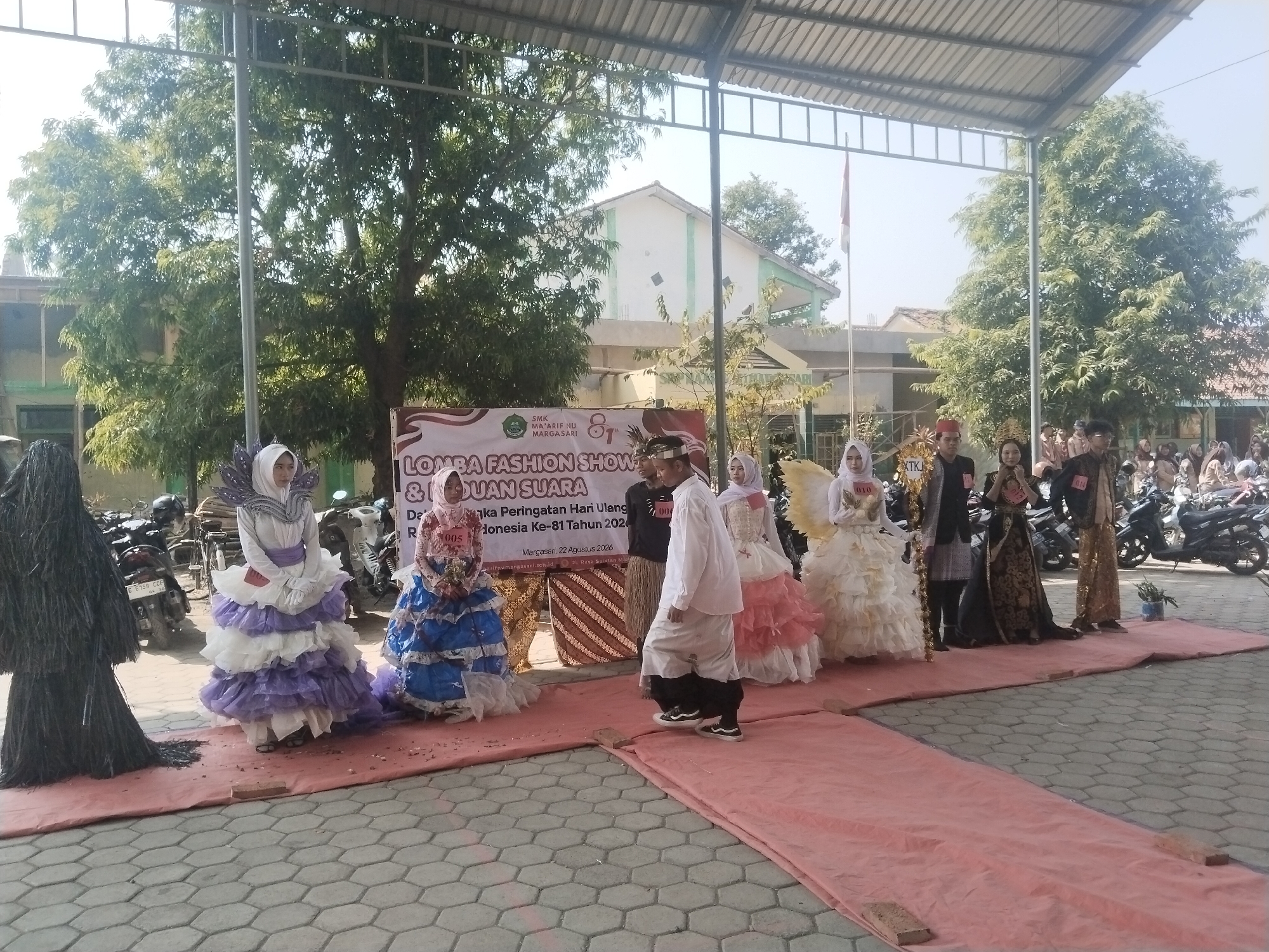 A group of individuals in elaborate outfits are posing for a fashion show on a red carpet laid out on a hexagonal paved surface. In the background, there is a banner that reads "LOMBA FASHION SHOW & DUAN SUARA" and "SMK MA'ARIEF NU MARGASARI". The event appears to be a school fashion show celebrating Indonesia's 81st anniversary, held on August 22, 2026. The location is identified as Margasan, Pakulaut, Indonesia. There are parked motorcycles behind the participants, and trees and buildings are visible in the distance. The participants are diverse, with some wearing traditional Indonesian attire and others in creative, possibly recycled material outfits. The atmosphere seems festive, with an audience of spectators visible in the background on the right side of the image.