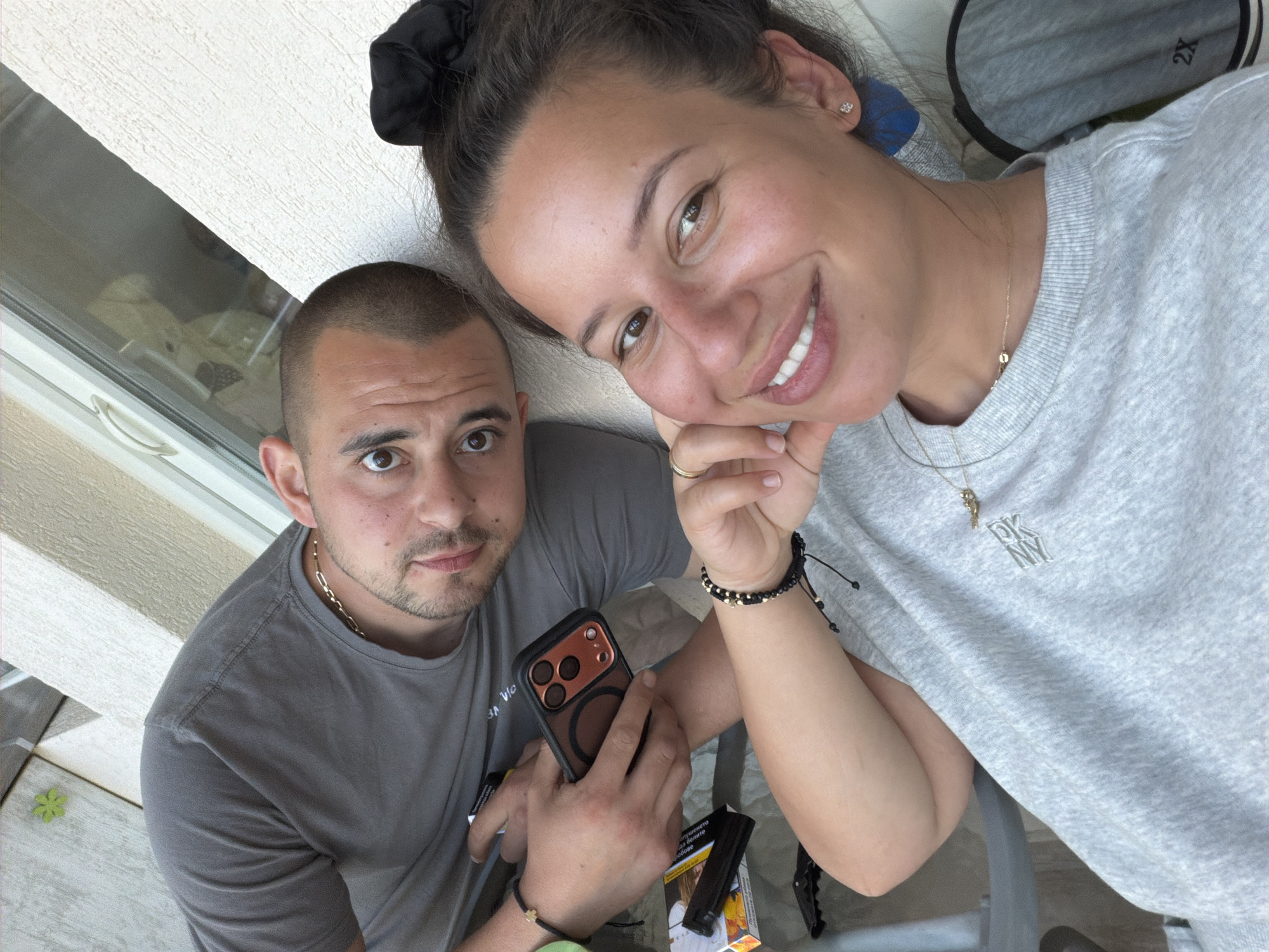 A couple is photographed outdoors, possibly on a balcony or terrace, with a building facade and window in the background. The man, on the left, is wearing a grey t-shirt and a gold chain necklace. He is holding a smartphone. The woman, on the right, is wearing a grey sweatshirt and a gold necklace. Both are looking at the camera. The scene appears to be in Burgas, Bulgaria.