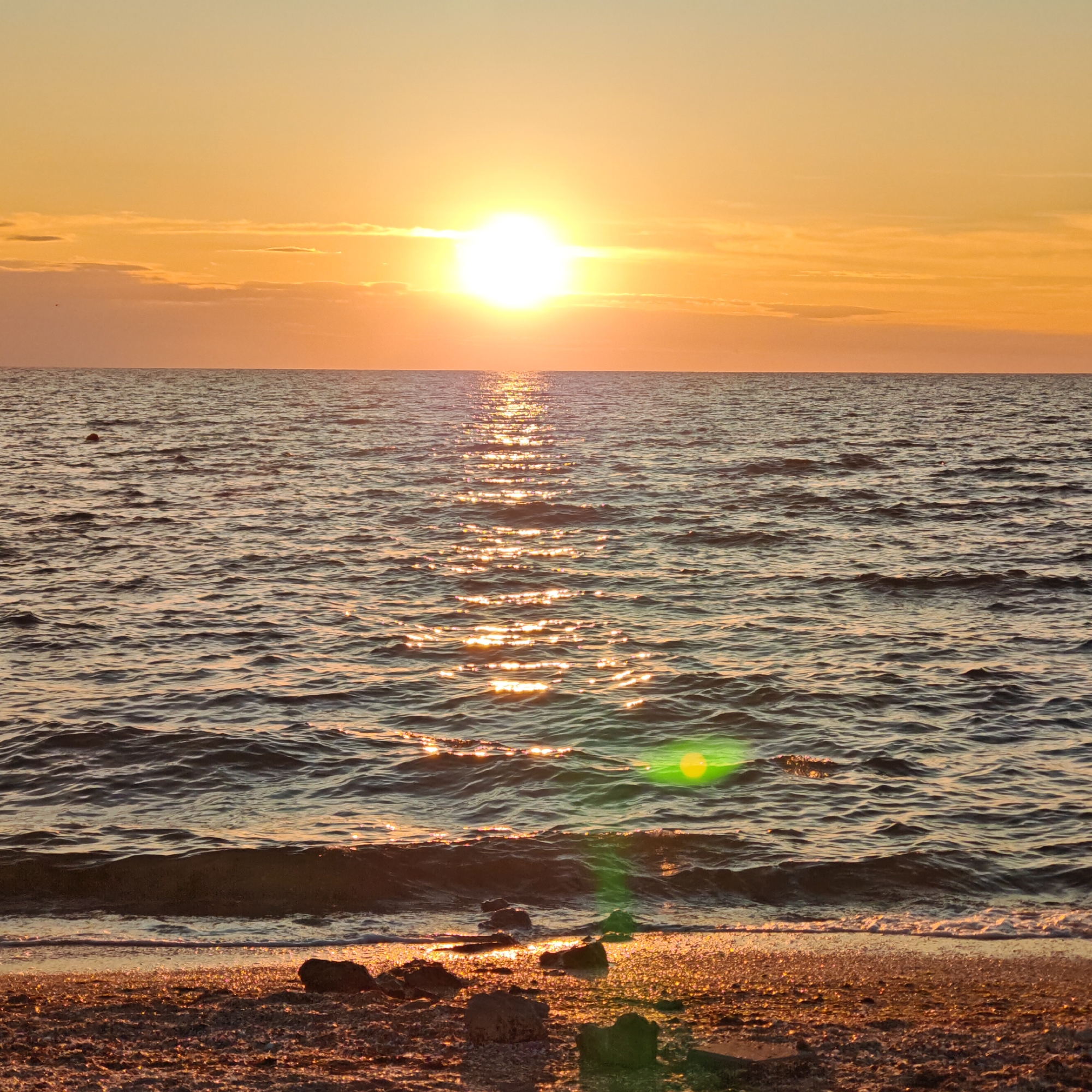 A scenic photograph captures a serene beach scene at sunset. The sun, a bright orb with a strong halo effect, hangs low in the sky, casting a warm golden glow across the water and sand. The horizon is a sharp line between the sky and the ocean, with wispy clouds tinged with orange and yellow. The sea is calm, with gentle ripples reflecting the sunlight, creating a shimmering pathway from the sun to the shore. On the beach in the foreground, pebbles and small rocks are scattered across the sand, glistening in the fading light. A faint green lens flare is visible in the lower right quadrant of the image. There are no people or animals in the frame.
