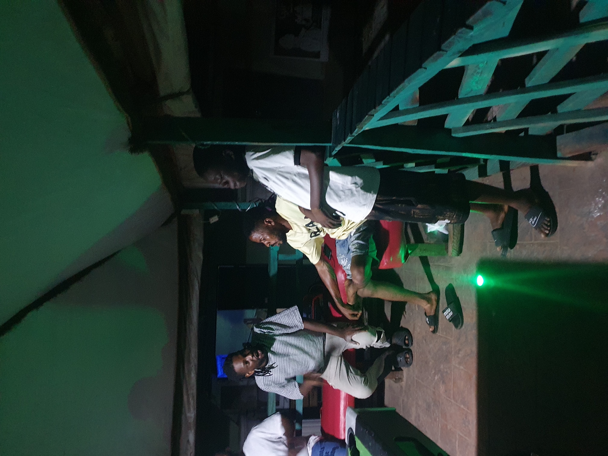 In Lokoja, Nigeria, four individuals are gathered in a dimly lit, open-air setting with a green ceiling. Three young men are seated on red benches. The man on the left, wearing a checkered shirt and light-colored pants, is gesturing with his hands as if speaking. The man in the middle, in a yellow t-shirt and shorts, is looking towards the man on the left. The third man, wearing a white t-shirt and dark shorts, is standing slightly apart and looking to his right. A fourth individual, partially visible on the far left, is wearing a white t-shirt and appears to be seated. A green railing is visible in the foreground, and the floor is tiled. A bright green light emanates from the floor near the center of the image. The overall ambiance suggests a casual social gathering.