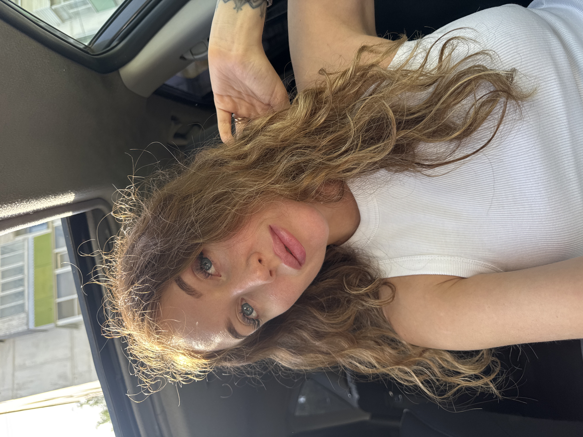 A woman with long, wavy brown hair and fair skin is in the driver's seat of a car, looking at the camera. She is wearing a white ribbed top. Her left hand is raised near the car's sunroof. There are buildings visible through the car window on the left. The image was taken in the city of Chaikovsky, Russia.