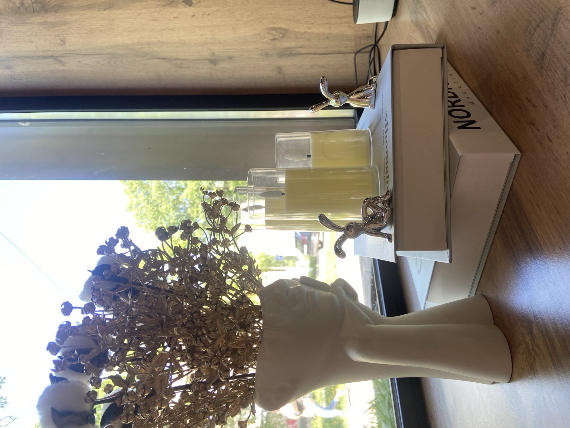 The image shows a still life arrangement on a wooden surface, likely a table or countertop. The arrangement consists of a white, human-shaped vase filled with golden floral decorations and cotton balls. Beside the vase are three cylindrical candles in glass holders, placed on top of two stacked white boxes. The top box has the word "NORDIC" printed on it. Two silver rabbit figurines are placed on the boxes. The scene is located near a window with a view of trees and a road, suggesting an indoor setting. The location within the city of Chaikovsky, Russia, cannot be definitively determined from the visual information provided.
