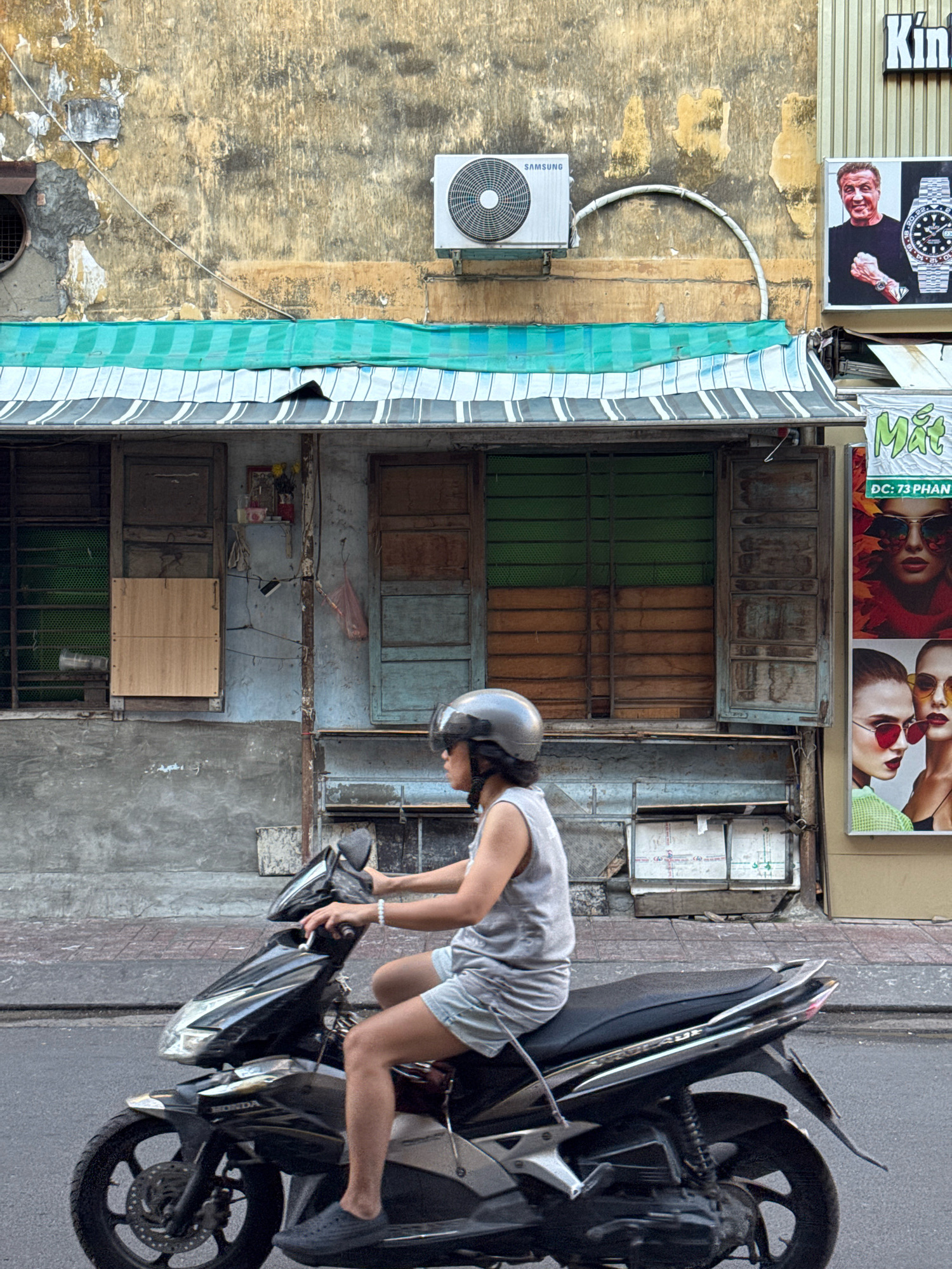 In Nha Trang, Vietnam, a person wearing a grey tank top, light blue shorts, a grey helmet, and dark grey slip-on shoes rides a black motorcycle down a paved street. The person is positioned in the center of the frame, on the left side of the motorcycle, facing right. The motorcycle is black and silver with a black seat and appears to be in motion. The background features a weathered, textured wall with peeling paint in shades of beige and grey. An air conditioning unit is mounted high on the wall, and a green and white striped awning stretches across a section of the building. A large advertisement featuring two women in sunglasses is visible to the right. The overall environment suggests an urban setting with elements of aging infrastructure and commercial advertising.