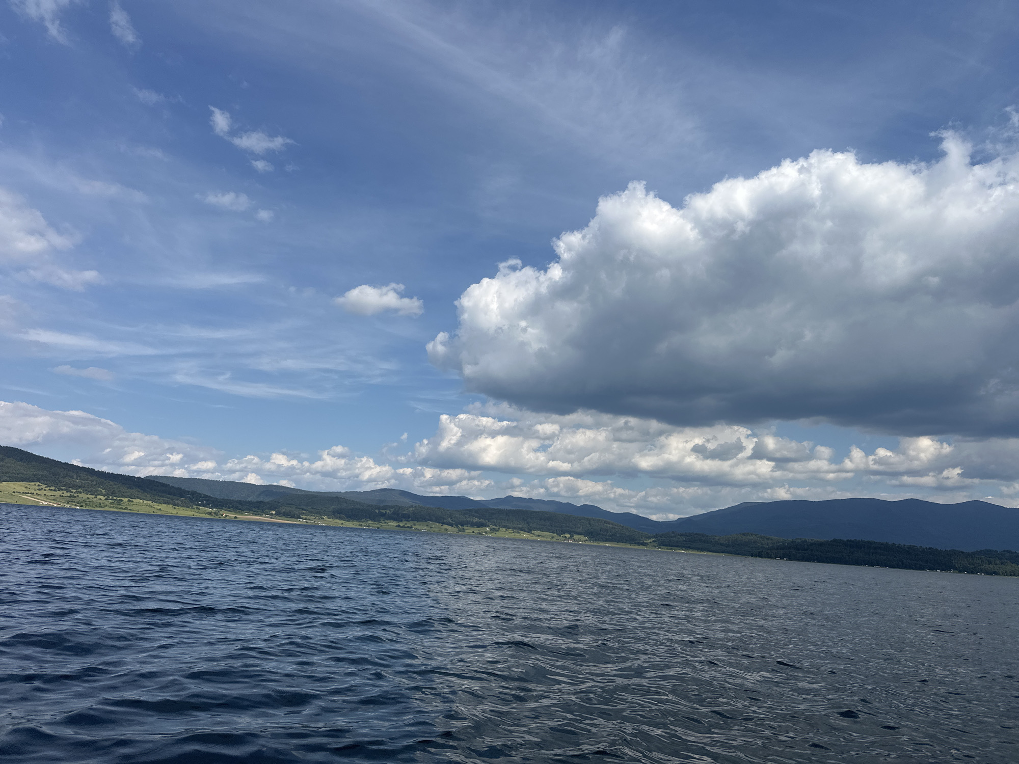 A wide-angle, eye-level shot captures a serene lake scene under a partly cloudy blue sky. The foreground is dominated by the dark blue, rippling surface of a large body of water, reflecting the sky above. In the mid-ground and background, a lush green shoreline stretches out, dotted with scattered trees and some buildings. Beyond the shoreline, rolling hills and distant mountains form a majestic backdrop. The sky is a vibrant blue, streaked with wispy cirrus clouds and punctuated by larger, fluffy cumulus clouds, particularly on the right side of the frame. The overall impression is one of natural beauty and tranquility. This scene is likely located in Batak, Bulgaria, given the context of the image.