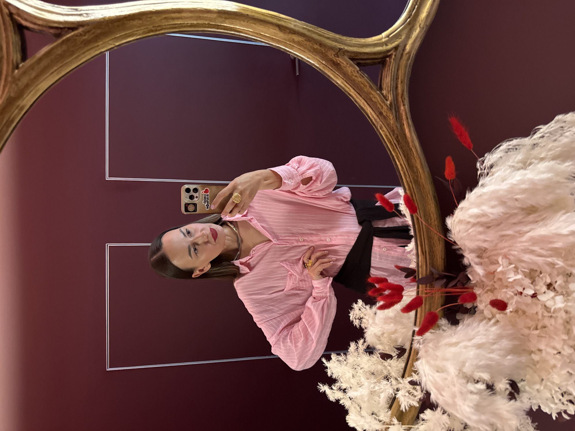 An indoor, mirror selfie captures a woman posing in front of a dark reddish-purple wall, framed by an ornate gold mirror. The image is rotated 90 degrees clockwise.
The subject is a woman with dark hair, wearing a light pink, striped button-up shirt with rolled-up sleeves and a black sash or belt. She has red lipstick and is adorned with gold jewelry, including a prominent chunky ring and a thick necklace. She holds a smartphone with a clear case that features a red heart (or cherry) graphic and Cyrillic text, which appears to read "Я люблю свой город" (I love my city) and "SOCHI".
The woman is taking a mirror selfie, looking directly at the camera with a neutral to slightly smiling expression. Her pose is casual but deliberate.
The setting appears to be an interior space, possibly a stylish room or boutique, suggested by the decorative elements. To the right, within the mirror's reflection, is a large, elaborate floral arrangement featuring fluffy white dried flowers (resembling pampas grass) and several bright red dried seed pods or flowers, adding a touch of vibrant contrast. The dark wall in the background has subtle rectangular outlines, suggesting architectural detailing. The overall ambiance is chic and well-decorated, with the context indicating the location is Sochi, Russia.