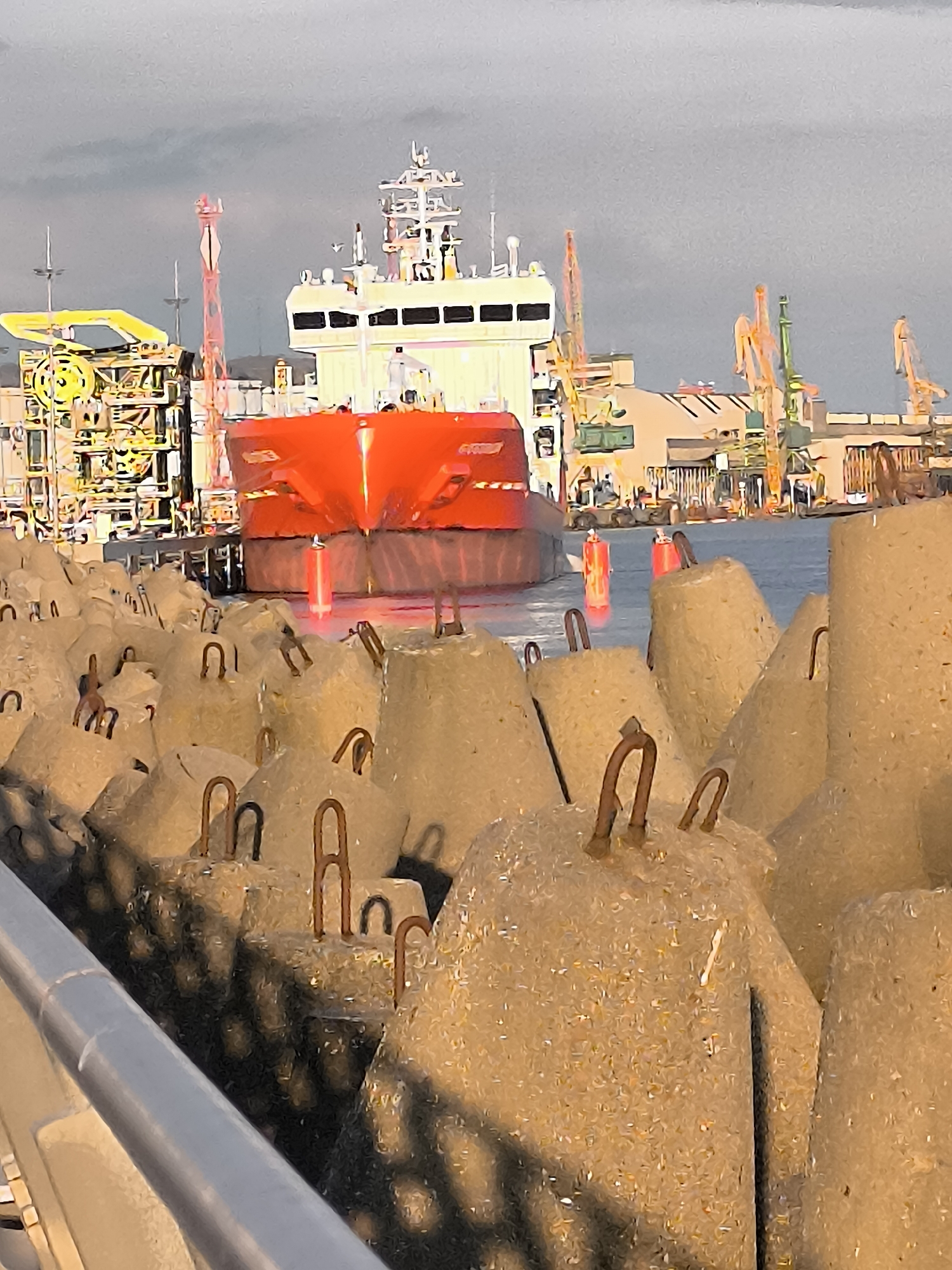 The image captures an industrial port or shipyard scene, dominated by the bow of a large, bright orange ship. The ship, with its white superstructure and bridge visible above the hull, appears to be docked or maneuvering within a harbor.
In the immediate foreground, a dense arrangement of rough-textured concrete blocks, likely breakwater units, fills the lower half of the frame. These blocks, in varying sizes and shades of beige and grey, feature rusty rebar loops protruding from their surfaces. A dark grey metal railing runs diagonally across the bottom left of the image.
Behind these foreground structures, a calm, dark body of water reflects muted light, with several red, cylindrical markers or buoys floating near the ship's hull.
The background reveals a bustling industrial environment. To the left of the ship, a complex yellow industrial structure stands tall, alongside a slender red crane or support tower. To the right and further back, numerous cranes, colored yellow and green, are visible, interspersed with industrial buildings and hangars. The sky overhead is uniformly overcast and light grey, suggesting a cloudy day or a muted light condition.
The overall aesthetic of the image appears somewhat stylized or digitally processed, lending a painterly quality with defined outlines and slightly flattened colors. While there are faint, blurry markings on the ship's hull and the yellow industrial structure, no decipherable text is visible. The scene conveys the functional and active atmosphere of a modern port under an overcast sky.