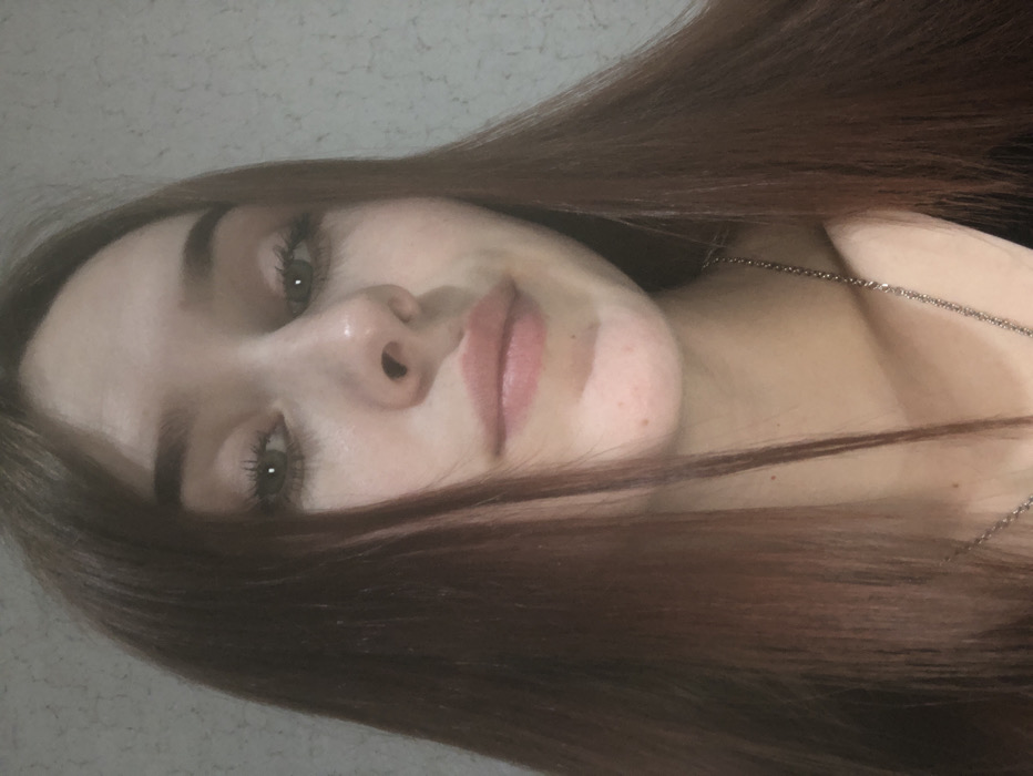 The image displays a head-and-shoulders portrait of a young adult female, taken indoors in the city of Фоки, Russia. She has long, straight, reddish-brown hair, parted near the center, which frames her face and extends over her shoulders. Her green or hazel eyes are open and directed at the camera. Her lips are closed, with a slight upward curve, indicating a subtle smile. Minor skin blemishes are visible on her chin. A thin, silver-colored chain necklace is worn around her neck, partially visible over a dark-colored garment. The background is a light-colored, textured wall. The photograph is taken from a slightly low angle.