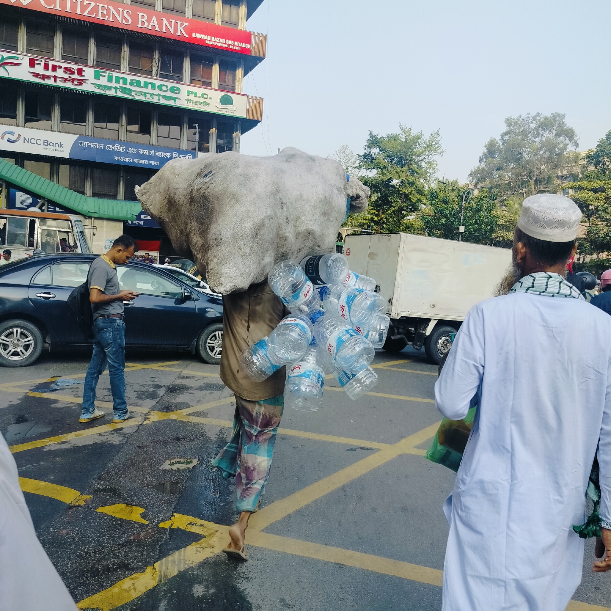 A man carries a large sack and numerous water bottles on his head in Dhaka, Bangladesh. He wears a lungi and walks barefoot on a street with yellow painted lines. Another man in a gray shirt and jeans looks at his phone near a dark blue car. A man wearing a white tunic and a cap walks in the opposite direction. In the background, a building with signs for Citizens Bank, First Finance PLC, and NCC Bank is visible. A white truck is parked on the side of the road. The image appears to be taken in the Kawran Bazar area of Dhaka.