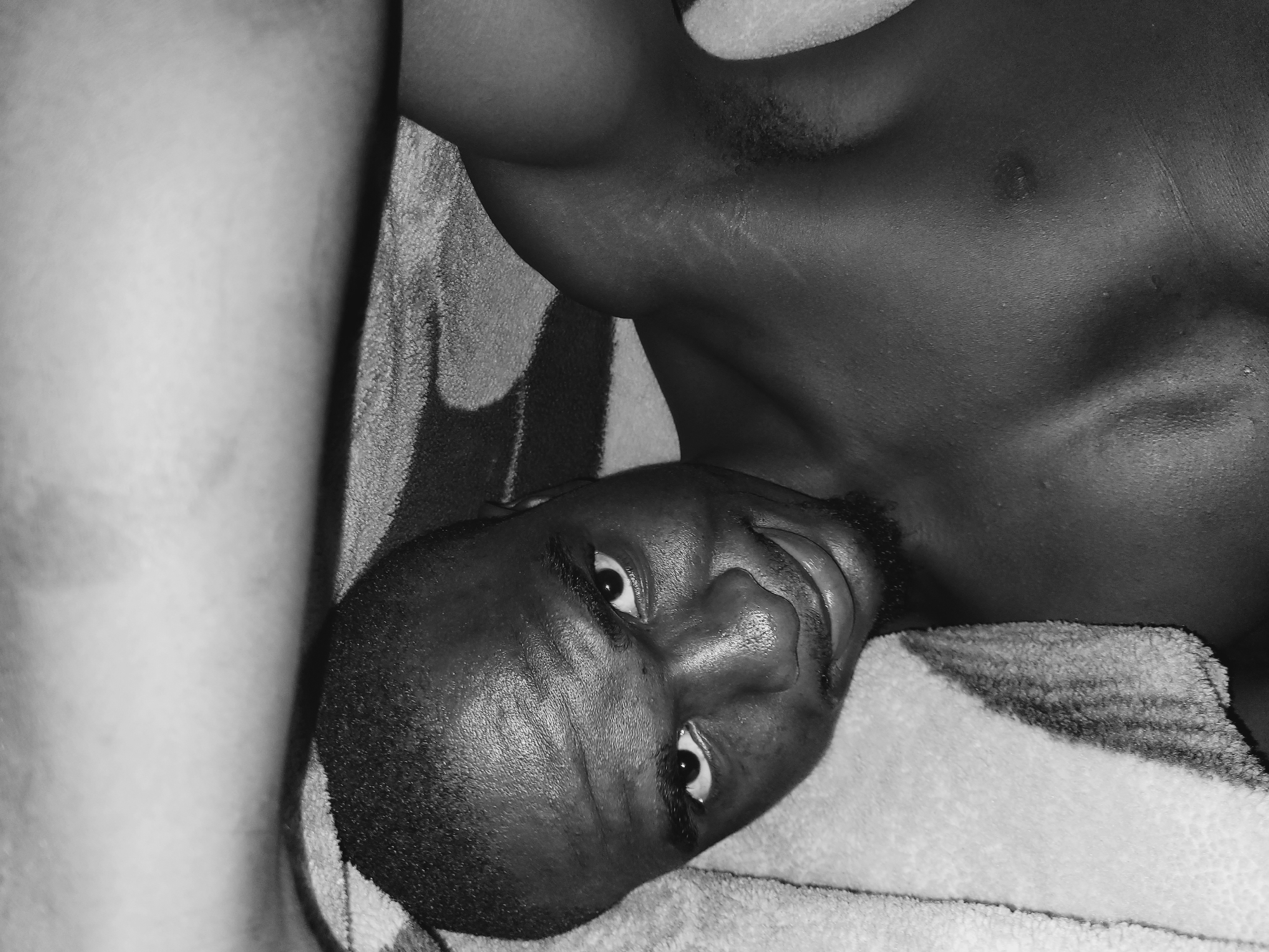 A grayscale, close-up, high-angle shot shows a Black man lying down. He is looking at the camera with a slight smile. His eyes are open, and his eyebrows are raised. He has a short haircut and a goatee. A light-colored towel is draped over the top of the image and partially covers his head and chest. His right arm is bent upwards and rests near his head, with his bicep visible. The background appears to be a patterned blanket. The image is taken in Bukuru, Nigeria.