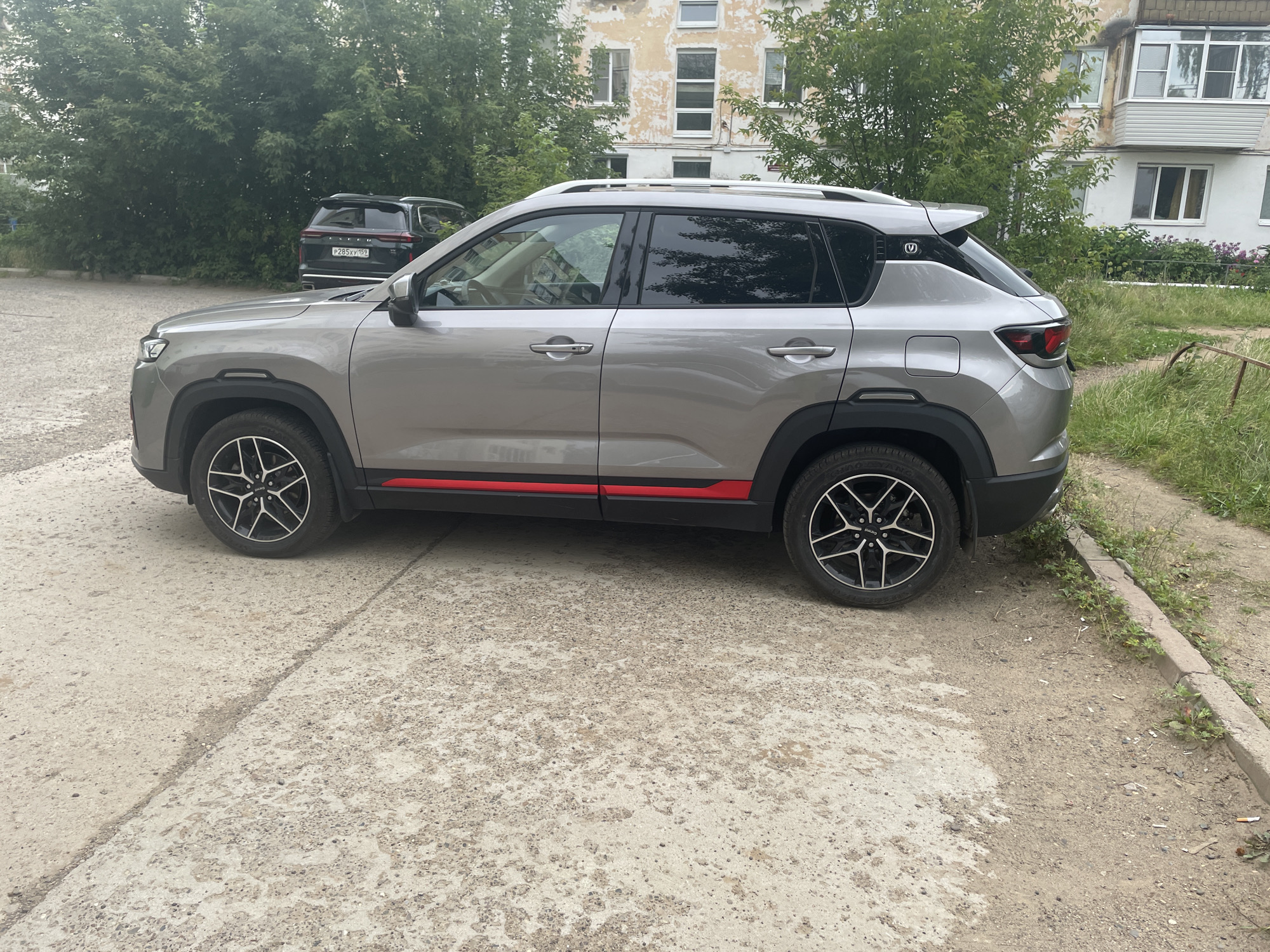 A grey, four-door SUV is parked diagonally on a cracked concrete surface. The car has black trim around the wheel wells and red accent stripes along the lower body panels. The rear wheels are silver and black with a multi-spoke design. Behind the SUV, a dark-colored SUV is visible, also parked. In the background, a weathered, light-colored apartment building stands with green trees and vegetation surrounding it. The scene appears to be an outdoor parking area in a residential district of Chaikovsky, Russia, likely on a cloudy day.