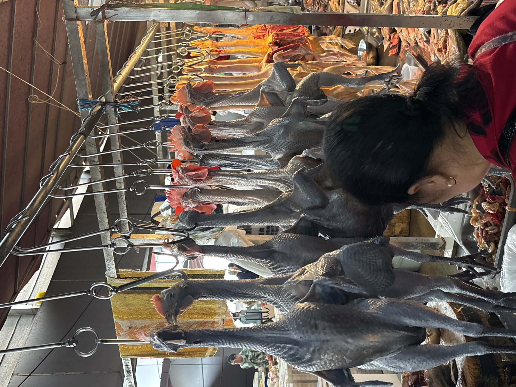 A woman in a red jacket examines a row of skinned chickens hanging from metal hooks in a market stall. The chickens have dark gray skin and are arranged vertically, with some paler fowl above. The location is Jiangjin, China, as reported from the internet.