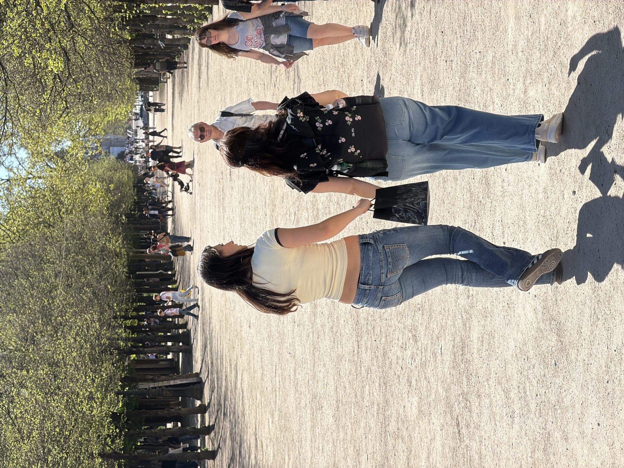 The image shows a sunny outdoor scene in Paris, France. A tree-lined promenade is visible, with benches and a pathway. Several people are present, including three women in the foreground. One woman is wearing a light yellow top and blue jeans. Another woman is wearing a floral top and blue jeans and is carrying a black bag. A third woman wears sunglasses and is dressed in a denim skirt and top. In the background, other people are walking along the path. The location appears to be a park in Paris.