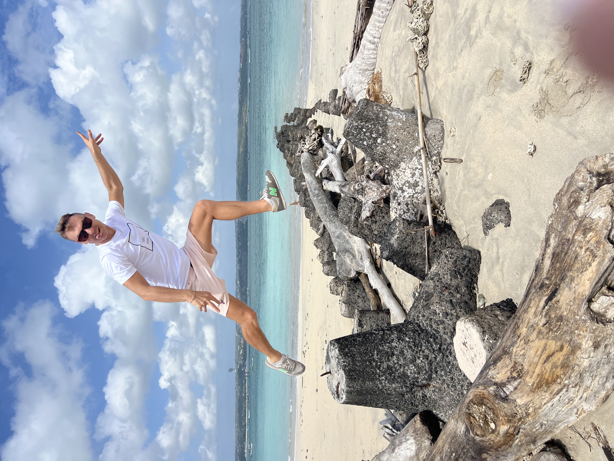 A man in a white t-shirt and pink shorts is captured mid-air, jumping over a barrier of rocks and driftwood on a beach. The man is wearing sunglasses and has his tongue sticking out, with his left arm extended to his right. His right leg is bent at the knee, and his left leg is extended back. The beach is sandy, with the turquoise ocean and a hazy coastline in the background. The sky is blue with scattered white clouds. This scene is likely on one of Denpasar's beaches, such as Pantai Padang Padang or Pantai Dreamland, known for their rocky formations and vibrant waters.