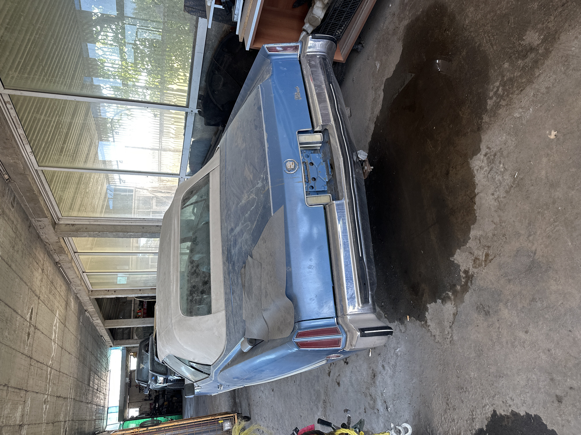 An abandoned blue Cadillac Eldorado convertible is parked in a dimly lit garage. The car is covered in dust and has a torn black tarp draped over its rear. The convertible top is also dusty. A large dark puddle of liquid is on the concrete floor beneath the car's rear. In the background, other vehicles and storage shelves are visible. The garage has a rough concrete ceiling and walls, with large windows that allow sunlight to filter in.
