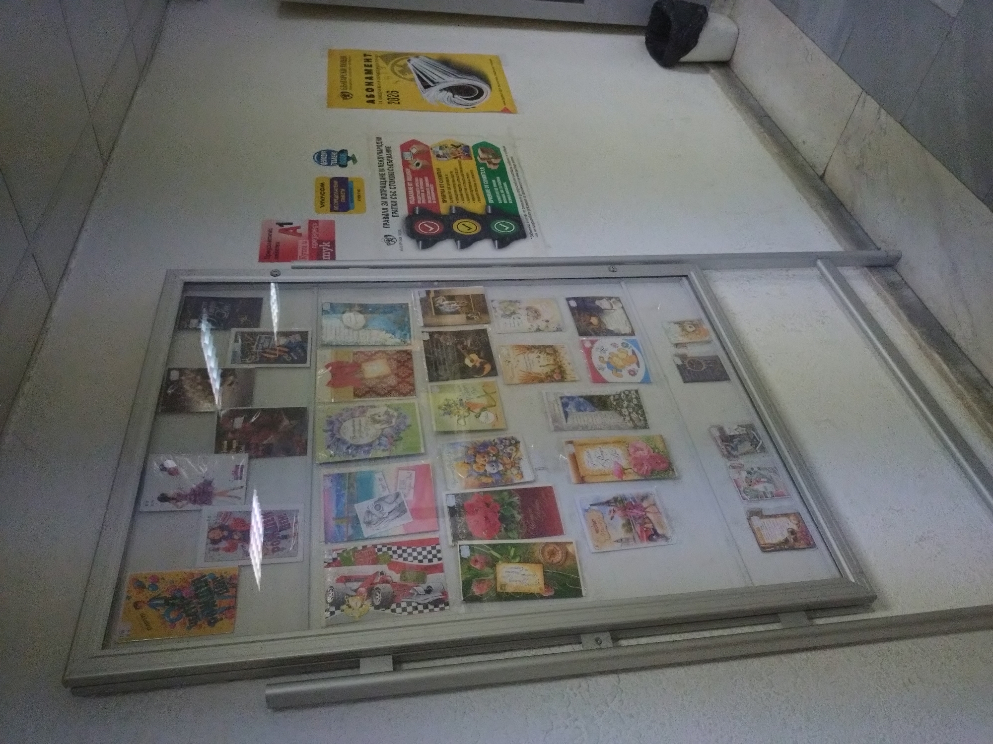 The image shows a display case containing various postcards, affixed to a wall. Above the display case are posters or signs, including advertisements for telecom providers A1 and Vivacom, as well as a sign related to international postal regulations. One poster features the word "ABOHAMEHT" and the number "2026". The wall is white, and the lighting appears to be artificial. The scene seems to be inside a public building, possibly a post office or a similar establishment. There's a dark, cylindrical object attached to the wall near the top-right corner of the image. The context of Stara Zagora, Bulgaria is only derived from the OCR text extracted from the signs in the image.