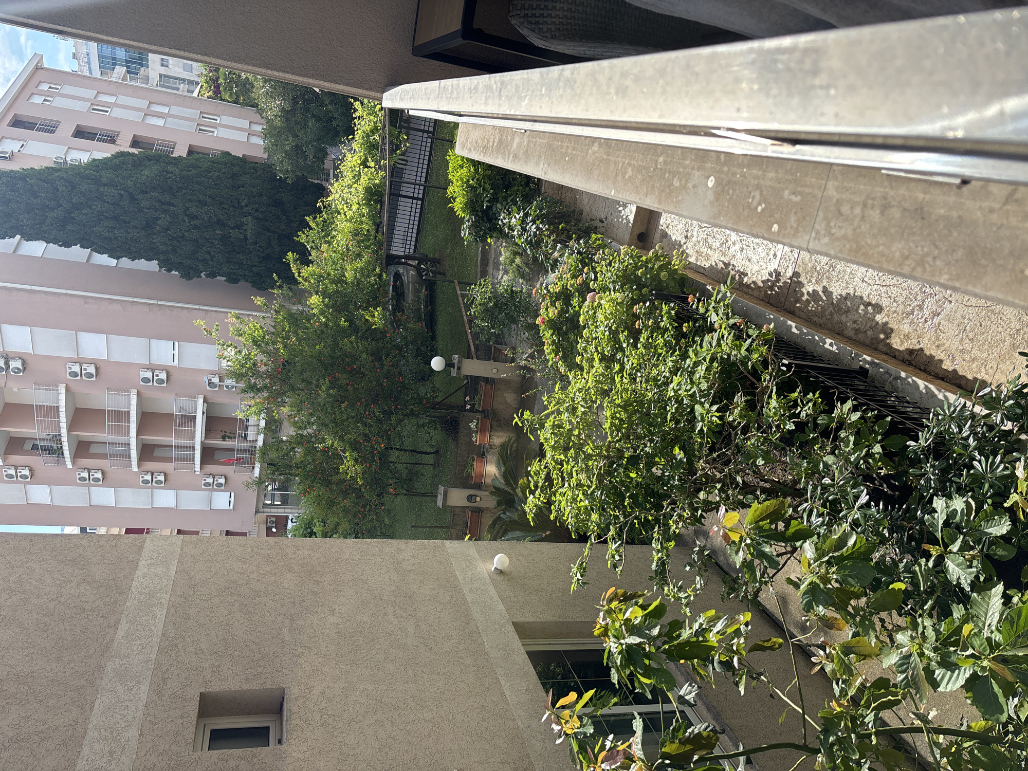 View from a balcony in Budva, Montenegro, looking down at a courtyard with trees and vegetation. In the background is a multi-story building with air conditioning units. The balcony railing is visible in the foreground. A section of a building is seen to the right and below with a window.