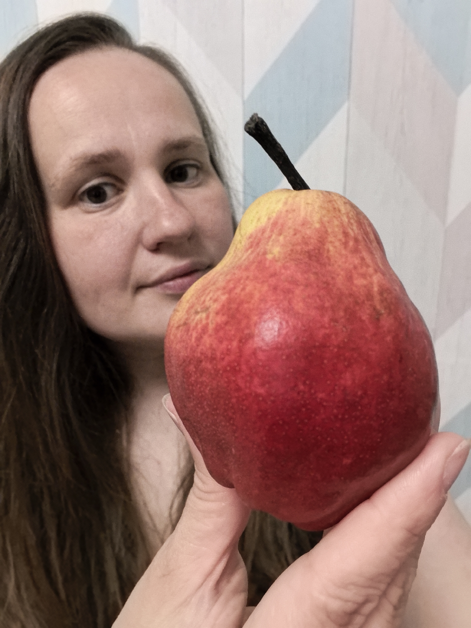 A woman is holding a red and yellow pear in front of a wall with a geometric pattern. The woman has long brown hair, fair skin, and is looking at the camera. The pear is the focal point of the image, with the woman's face slightly out of focus in the background. The location, Фролы, Russia, is not identifiable within the image.
