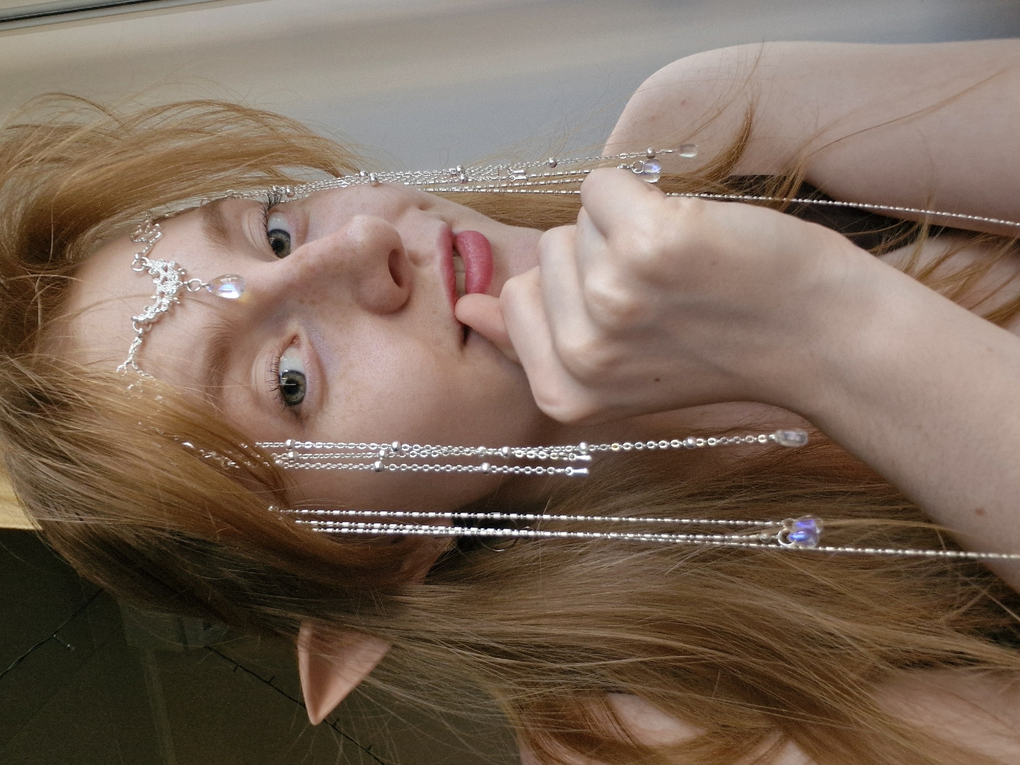 A fair-skinned individual with long, light brown hair is seen in a close-up shot, with the image oriented on its side. The person is adorned with a silver headdress featuring delicate chains and gemstone-like pendants. Their ear is pointed, suggesting an elven or fantastical theme. The background appears to be a simple, light-colored surface. The person's right hand is raised, with a finger near their lips. There is no indication of this image being taken in Yekaterinburg, Russia.