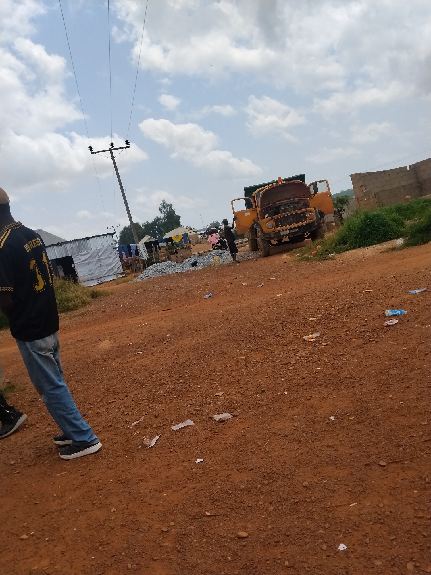 In Durumi, Nigeria, a dirt road is visible with a large yellow dump truck parked in the center of the frame. The truck has its hood and doors open. To the left of the truck, a person wearing a black and gold shirt with the number "31" on it and blue jeans stands facing away from the camera. The background shows a cloudy sky, some buildings with corrugated metal roofs, a pile of gravel, and lush green vegetation. Several plastic bottles and pieces of trash are scattered on the ground.