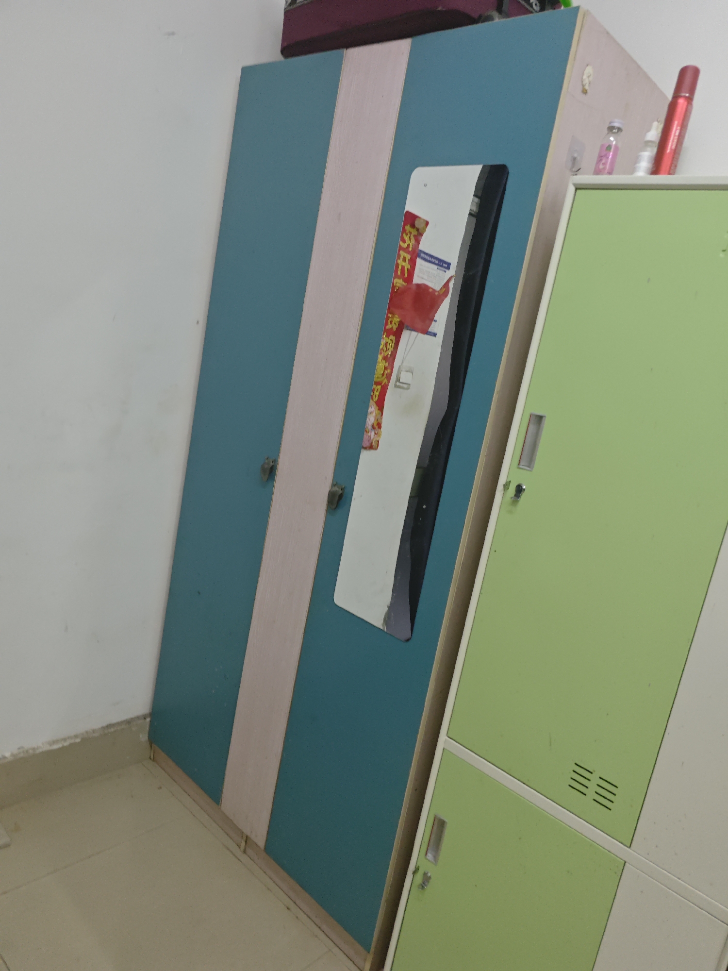 In an indoor setting, a large, blue and pink striped wardrobe stands diagonally against a white wall. A rectangular mirror is inset into the wardrobe, reflecting a red object, possibly a marker or pen, and two small bottles. To the right of the wardrobe, a light green locker with multiple compartments is visible. The floor appears to be tiled in a neutral color. The scene is static and appears to be a domestic or storage area. There are no individuals present in the image.