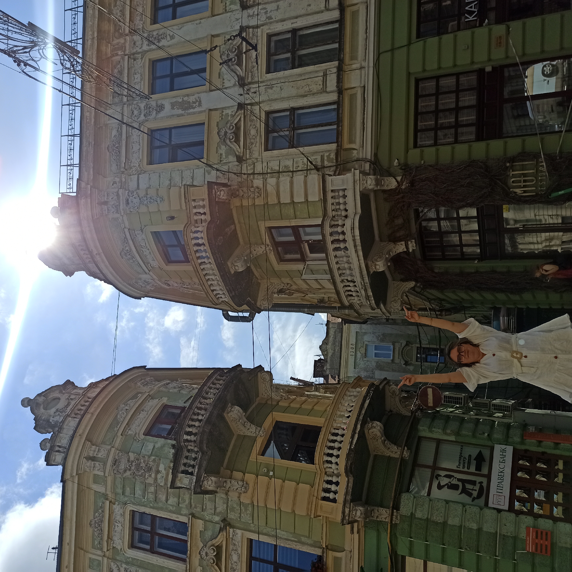 The image depicts a street scene in Chernivtsi, Ukraine, with a focus on two ornate, aged buildings with balconies and intricate architectural details. The buildings appear to be located in a historical district, evidenced by their classical style and weathered facades. The sky is bright blue with some clouds, and the sun is strongly visible, creating a lens flare effect.
In the foreground, a woman wearing a white dress and glasses is standing with her arms raised and fingers pointed upwards. She is positioned in front of the entrance to one of the buildings, which has a dark green lower section and large windows. A sign above a doorway reads "ПРАВЕКС БАНК" in Ukrainian. Another sign for "Motti Restauran" is visible on the right building. Overhead, a tangle of electrical wires crisscrosses the frame, typical of urban environments. In the background, other buildings are visible, suggesting a dense urban setting. One building in the distance has the inscription "1908". The overall impression is one of a sunny day in a city with rich architectural heritage.