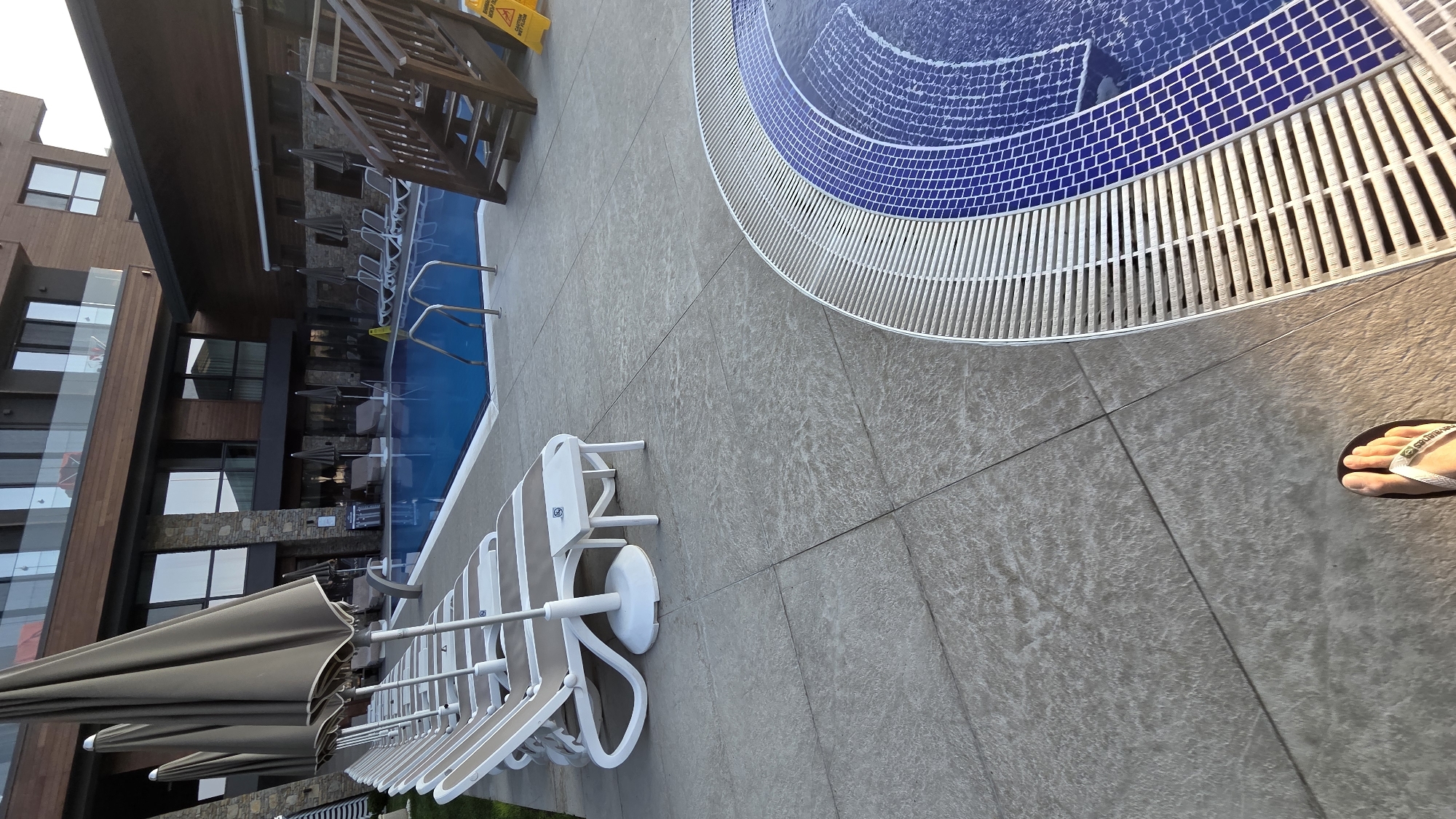 This media file captures an outdoor scene featuring a swimming pool area adjacent to a building. The focus is on the pool itself, which has blue tiled bottom with a circular pattern, and a white slatted edge. Several lounge chairs with light-colored fabric and white frames are arranged on the tiled patio surrounding the pool. Some of these chairs are folded and stacked, and one is partially open. Several closed umbrellas are also visible, suggesting they are stored.
To the left of the pool, a multi-story building with a modern architectural style is visible. It has a dark wood-paneled exterior and large windows with glass railings. The building appears to be a hotel or resort. The ground is paved with large, light-colored tiles that have a slightly textured, stone-like appearance.
In the lower right corner, a person's foot wearing a flip-flop is visible, stepping onto the tiled ground. The lighting suggests it is daytime, possibly late afternoon, given the warm tones of the light. The weather appears to be clear. The overall impression is one of a relaxed resort or hotel environment, likely in a warm climate, designed for leisure and swimming. There is a yellow caution sign with black text visible in the top center portion of the image, though the text is not clearly legible.