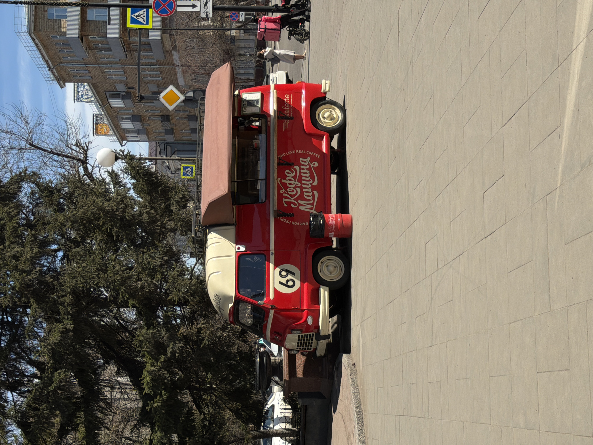 A vibrant red and cream vintage-style coffee truck, labeled "Кофе Машина" (Coffee Machine), stands prominently on a paved sidewalk in Krasnoyarsk, Russia. The truck, featuring the large number "69" on its side, also displays "WHO LOVE REAL COFFEE" and "BAR FOR PEOPLE," along with operating hours "4:30-1:00" and the text "Estafelke" or "etafette" as part of its branding.
The scene depicts a sunny daytime setting with a clear blue sky. Behind the coffee truck, a person can be seen walking on the sidewalk. To the left, a dense evergreen tree and a bare deciduous tree partially obscure a multi-story building. The building has a brick-like facade with numerous windows, some of which appear to have covers or decorations.
Several traffic signs are visible in the background, including a blue square sign with a yellow pedestrian crossing symbol, a red circular "no entry" or "no stopping" sign, and a yellow diamond-shaped "priority road" sign, alongside a white sign with a black arrow. A lamppost stands near the trees. The sidewalk is made of light-colored pavers, and sunlight casts shadows, indicating bright weather. The overall impression is a lively urban street scene centered around the mobile coffee shop.