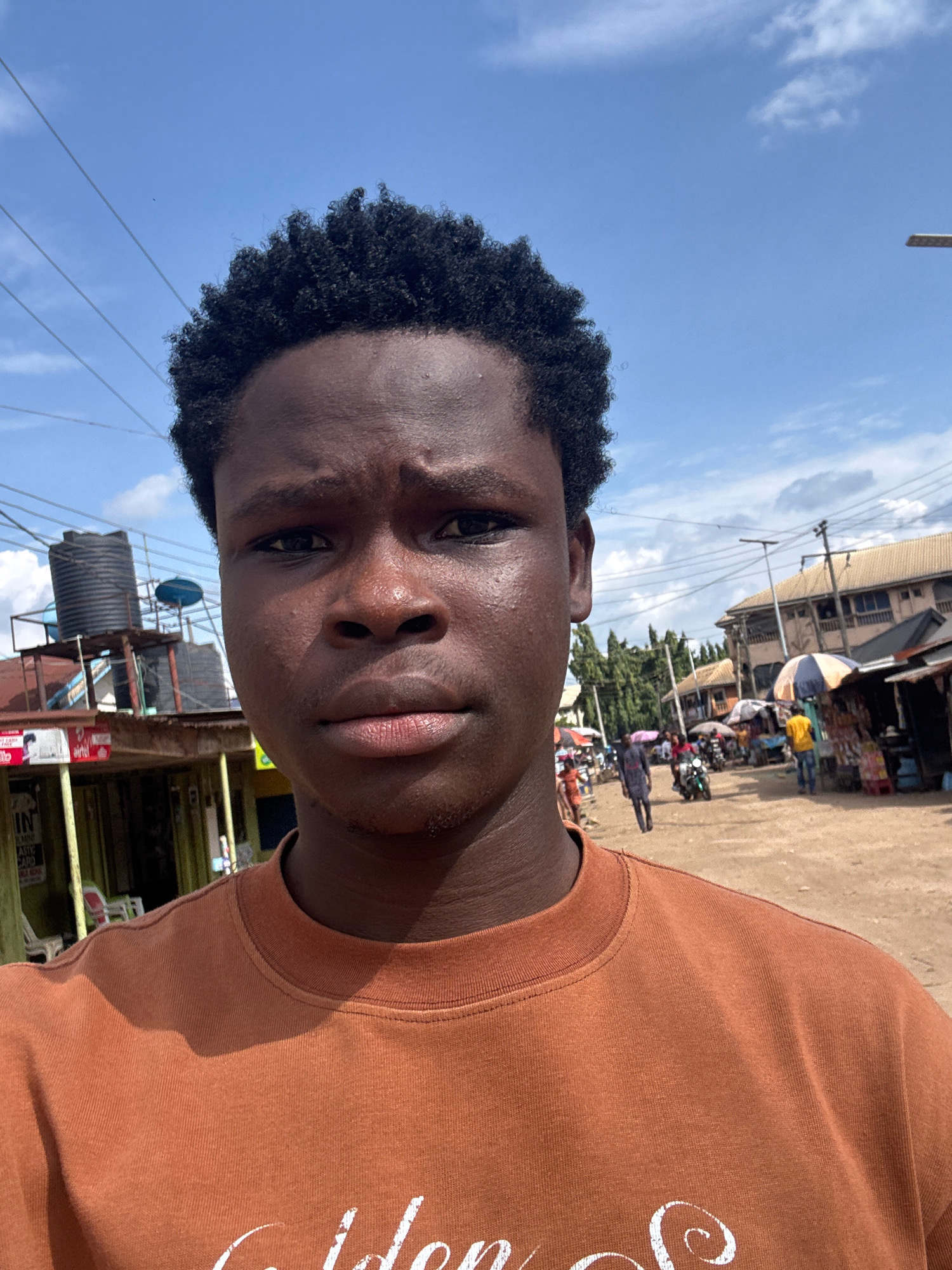 A selfie of a young Black male is taken outdoors in Ihiagwa, Nigeria. The subject is positioned in the foreground, facing the camera directly, with his head and shoulders filling most of the frame. He has short, dark, curly hair and is wearing a rust-colored crewneck sweatshirt. The background reveals a busy street scene with several small shops, market stalls, and various people walking and interacting. In the middle ground, there's a dirt road with a motorcycle and a person pushing it. Further in the background, multiple buildings with light-colored roofs are visible, along with numerous trees and utility poles. The sky above is bright blue with scattered white clouds. The overall atmosphere suggests a daytime setting in a bustling urban or semi-urban environment.