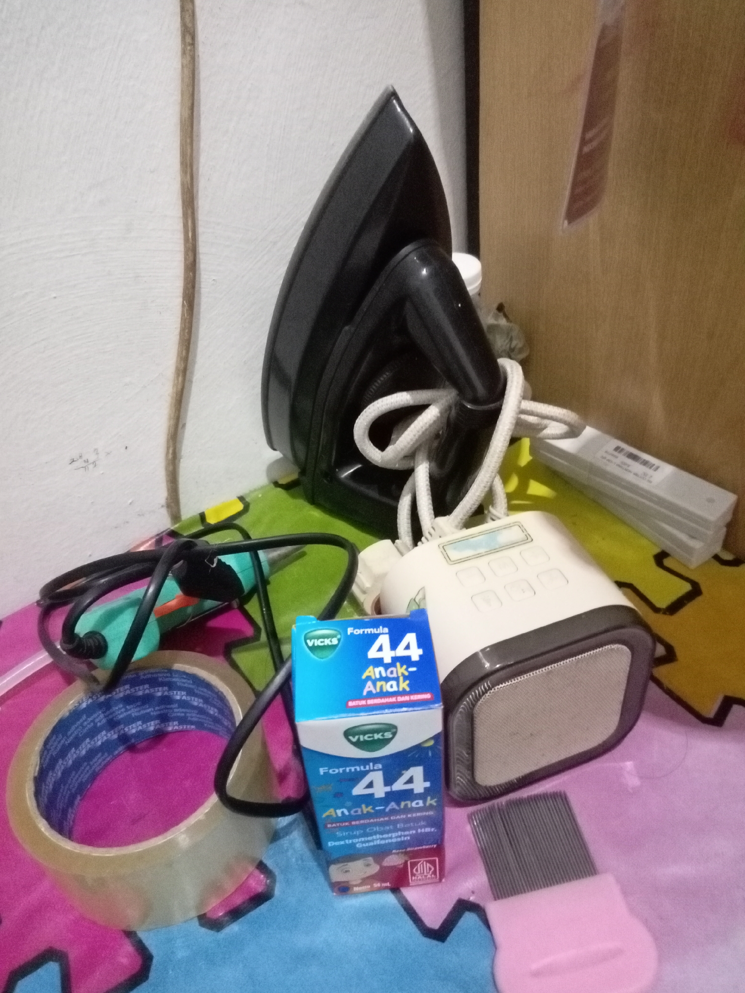 The image depicts a collection of household items on a patterned surface. Dominating the center-left is a black electric iron with its cord neatly wrapped around it. To the right of the iron is a white electronic device with buttons and a speaker grille. In front of these is a box of "Vicks Formula 44 Anak-Anak" cough medicine. To the left of the Vicks box, a roll of clear tape is visible. A green and black electric tool, possibly a soldering iron or glue gun, lies partially obscured by the tape and Vicks box. In the background, a stack of white rectangular objects, possibly test strips or similar medical supplies, are present. The overall environment appears to be indoors, with a wall and a partition in the background. The scene is likely set within a home or small storage area in Batam, Indonesia, given the presence of local products like the Vicks medicine for children.
