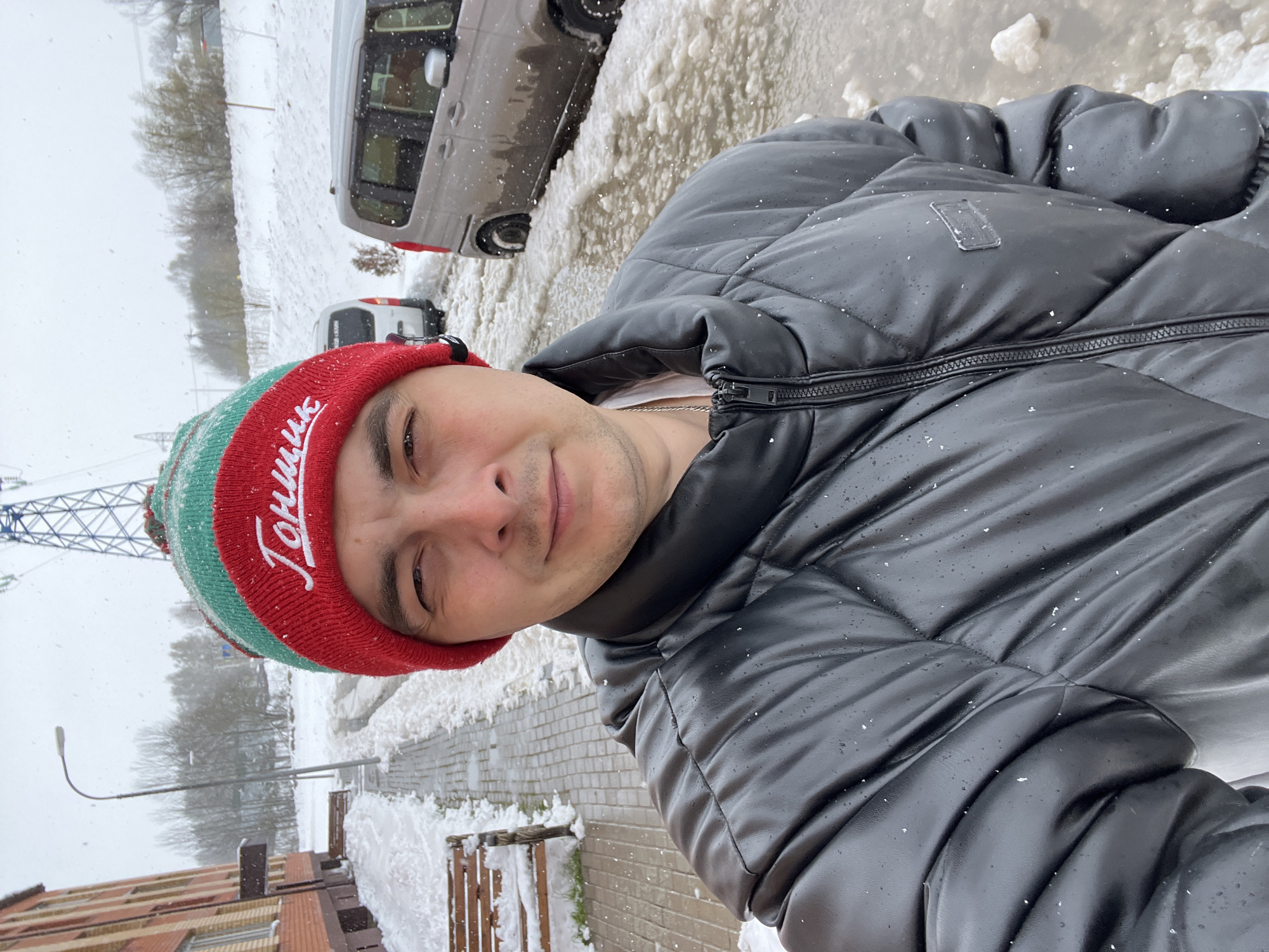 A male individual is captured in a vertical, eye-level selfie, looking directly at the camera with a subtle, content smile. He is wearing a red and green knitted winter hat with a white pom-pom, upon which the Cyrillic word "Тончик" (Tonchik) is embroidered in white text. He is also dressed in a black, possibly faux leather, puffer jacket with a visible zipper and a silver chain around his neck.
The setting is outdoors in what appears to be a snowy, urban environment, likely Vnukovo, Russia, given the context. Light snow is actively falling, visible as small white flakes scattered across his jacket and hat. The ground in the background is covered in a mixture of snow and slush. To the right of the individual, a grey minivan-like vehicle is parked, partially covered in snow. Further back and to the left, another white van-like vehicle is visible, also partially obscured by snow.
The background also features a paved sidewalk, brick buildings, and bare trees. A prominent power line or utility pole structure with intricate metallic supports is visible on the left side of the frame, along with a street light. The overall lighting suggests an overcast daytime sky, typical of winter weather. The scene conveys a calm, cold atmosphere with the individual seemingly embracing the snowy conditions.