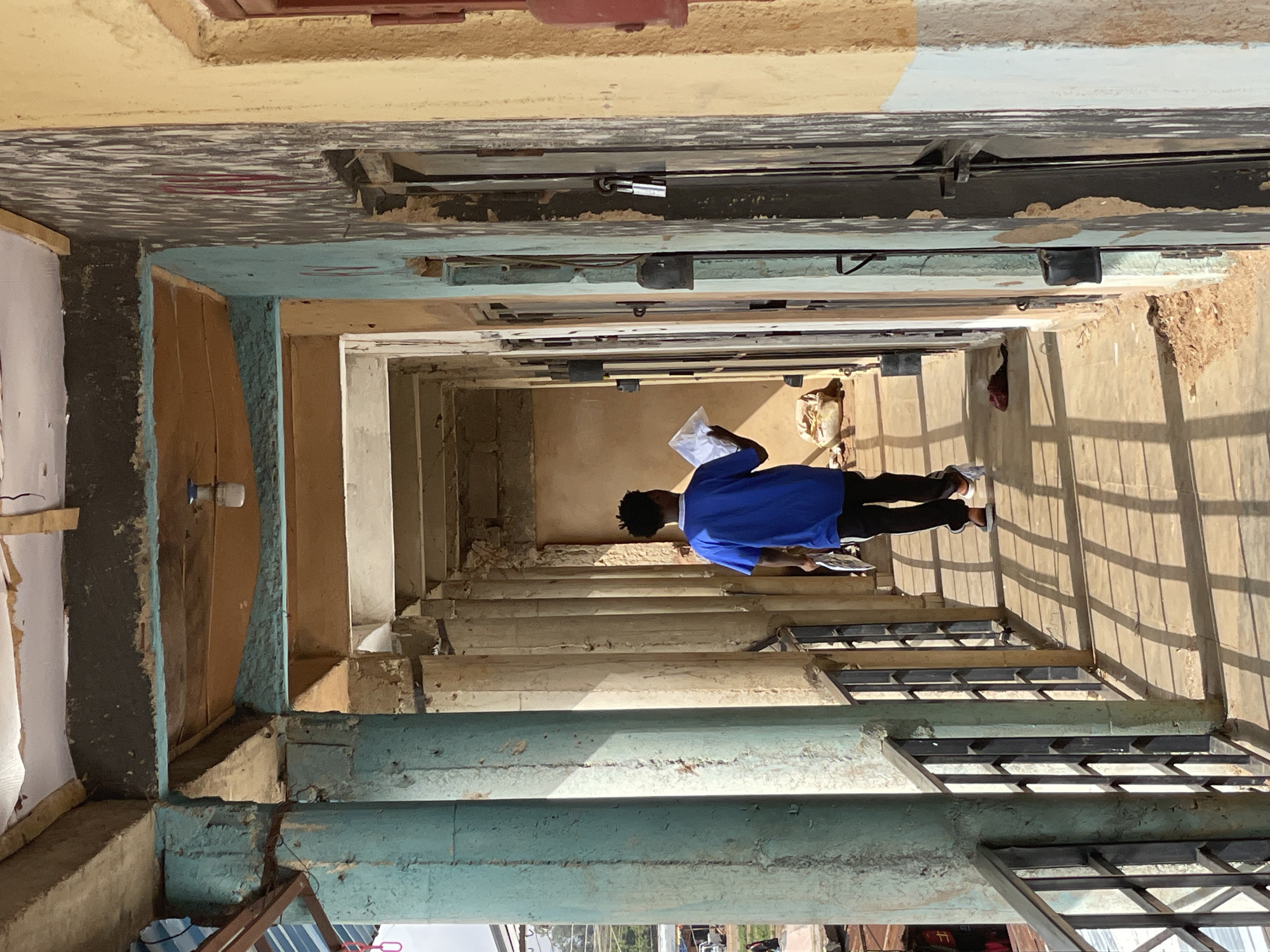 A person wearing a blue shirt and black pants is standing on a set of stairs in what appears to be an unfinished or dilapidated building in Bukuru, Nigeria. The person is holding papers. The building is constructed with concrete and metal features, and there are visible signs of decay or disrepair throughout the structure. The lighting suggests a sunny day. Outside of the structure, there is a view of trees, buildings, and vehicles.