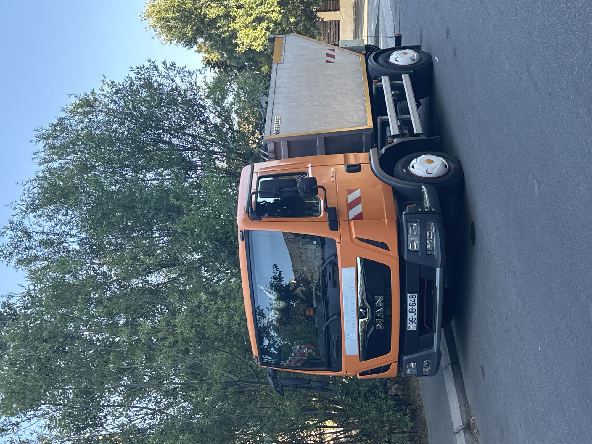 An orange MAN truck with a gray dumpster is parked on the side of a paved road in Baku, Azerbaijan, with a large tree and other foliage to the left and a building visible in the background. The truck's license plate reads "99-JB-648". The dumpster has a yellow stripe along its top edge and reflective red and white markings on its rear. The truck's cab has a banner with text and logos, and the windshield reflects trees and a building. The sky is clear and blue. The scene is captured during daylight.