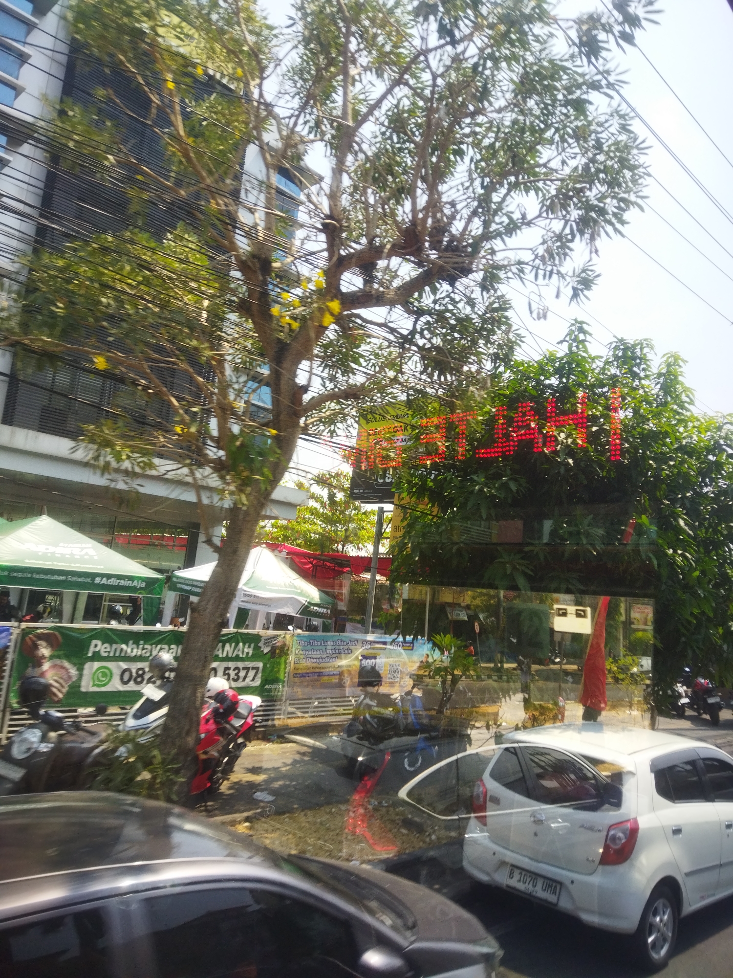 The image shows a street scene in Grogol, Indonesia. A tall, modern building with glass windows is on the left side of the frame. In front of the building, several green tents are set up, possibly for a promotional event. A tree with sparse foliage stands in the foreground, partially obscuring the view of the building and tents. Red, LED illuminated signage, displaying what appears to be Indonesian text, is visible behind the tree. Several motorcycles are parked on the side of the road, including a red and white motorcycle and a dark-colored one. A white car is parked in the foreground on the right side of the image, with its license plate clearly visible, reading "B 1070 UMA". The street is paved and appears to be a busy urban area. Reflections in the glass of the building show more of the street scene, including more vehicles and possibly pedestrians. The sky is clear and bright, indicating daytime.