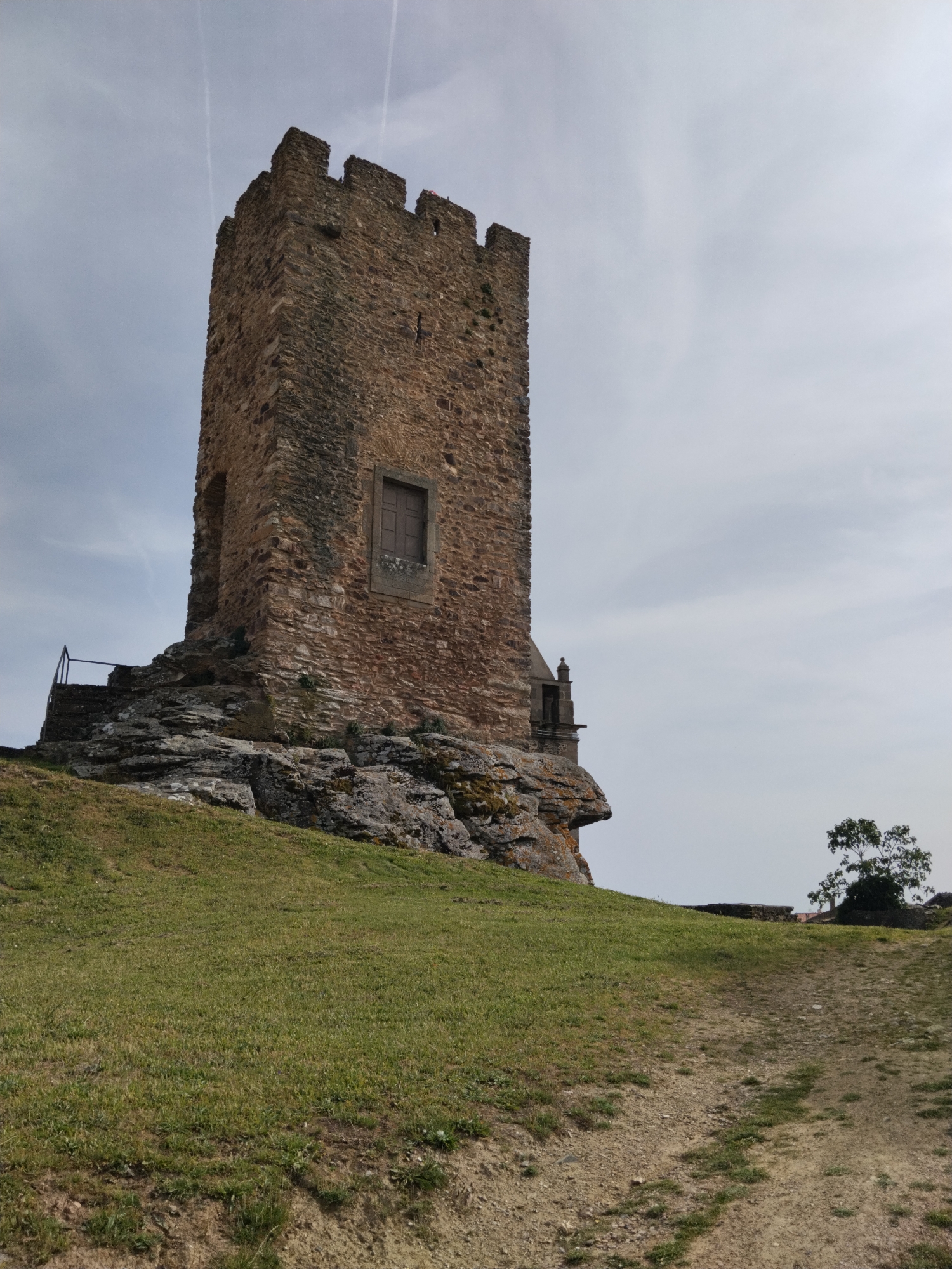 The image depicts the imposing tower of the Mogadouro Castle in Mogadouro, Portugal. The tower is constructed of brown stone, with crenellations along the top. The tower has a window on its facade. The tower sits on a rocky outcropping, accessible by stairs with a metal railing. The surrounding landscape is a grassy hill that slopes downwards to a dirt path. A tree stands to the right of the tower on the horizon. The sky is overcast.
