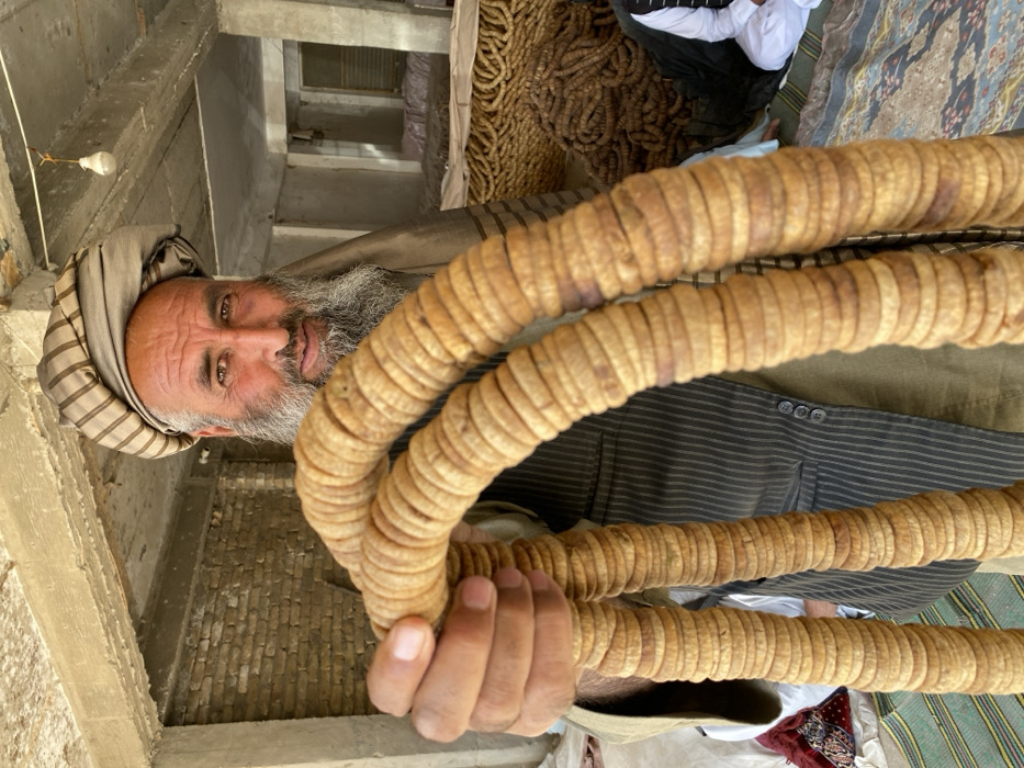 The image portrays an indoor commercial space, likely a market stall or storage facility, in Kandahar, Afghanistan. A male individual, appearing middle-aged with a full dark beard and a striped turban, occupies the central foreground, looking directly at the camera with a slight smile. He wears a dark pinstriped vest over a lighter tunic. The man holds two large, U-shaped strings of dried, flattened, light-brown figs prominently in front of him.
The immediate background on the left reveals an unfinished brick wall and concrete structural beams and columns. To the right, the interior features a concrete ceiling and support columns. A single bare incandescent lightbulb hangs from the ceiling. The floor on the right side of the frame is covered with patterned rugs and mats, upon which numerous coiled bundles of similar dried produce are stacked. A person, partially visible and clad in white, sits on a rug in the background to the far right. The floor on the left displays a green and white striped mat. The overall environment is rustic and functional, consistent with a facility for processing or selling dried goods.