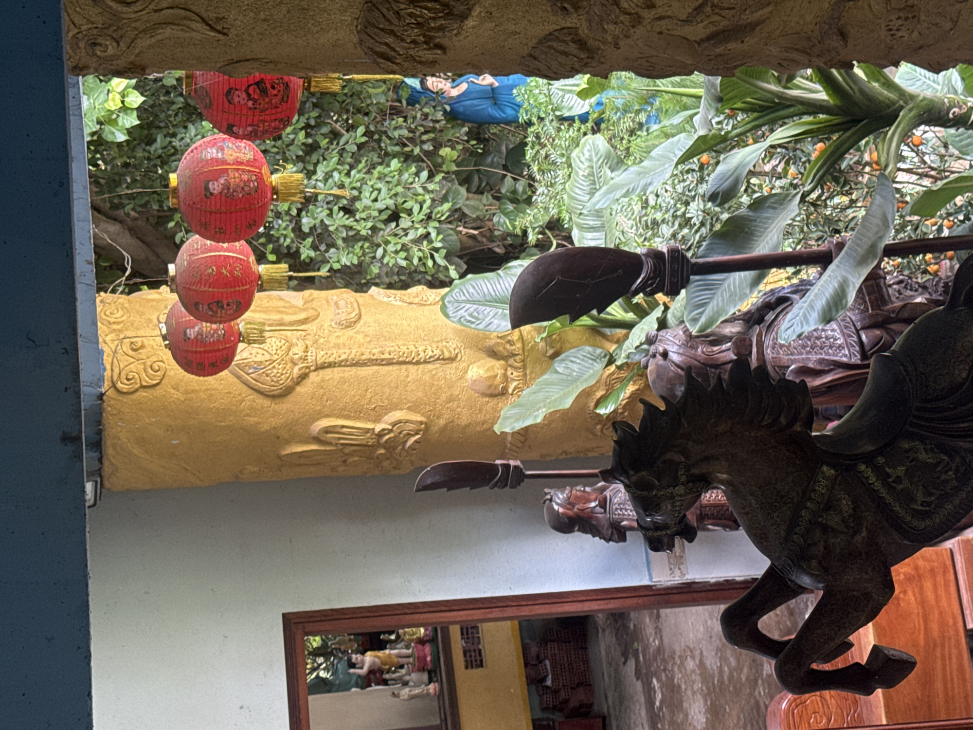 The image captures an outdoor scene, likely within the grounds of a religious or cultural site in Da Lat, Vietnam, given the architectural style and decorative elements. In the foreground, a dark, intricately carved wooden sculpture of a horse is prominent, its front legs raised as if in motion. To the left and slightly behind the horse, a wooden figure, possibly a warrior or deity, stands with a weapon.
Further back, beyond an ornate, textured yellow column, is a verdant garden area. Red Chinese lanterns with decorative tassels are suspended from above, adding a festive or traditional ambiance. Peeking through the foliage, a statue of a person in a blue robe is visible. The background suggests a lush, overgrown environment typical of Da Lat's climate.
To the left, a doorway or opening reveals an interior space with a mirror reflecting more religious statues and brickwork, indicating the presence of a building adjacent to the garden. The overall impression is one of a tranquil, culturally rich setting, possibly a temple or a decorative garden within a larger complex.
