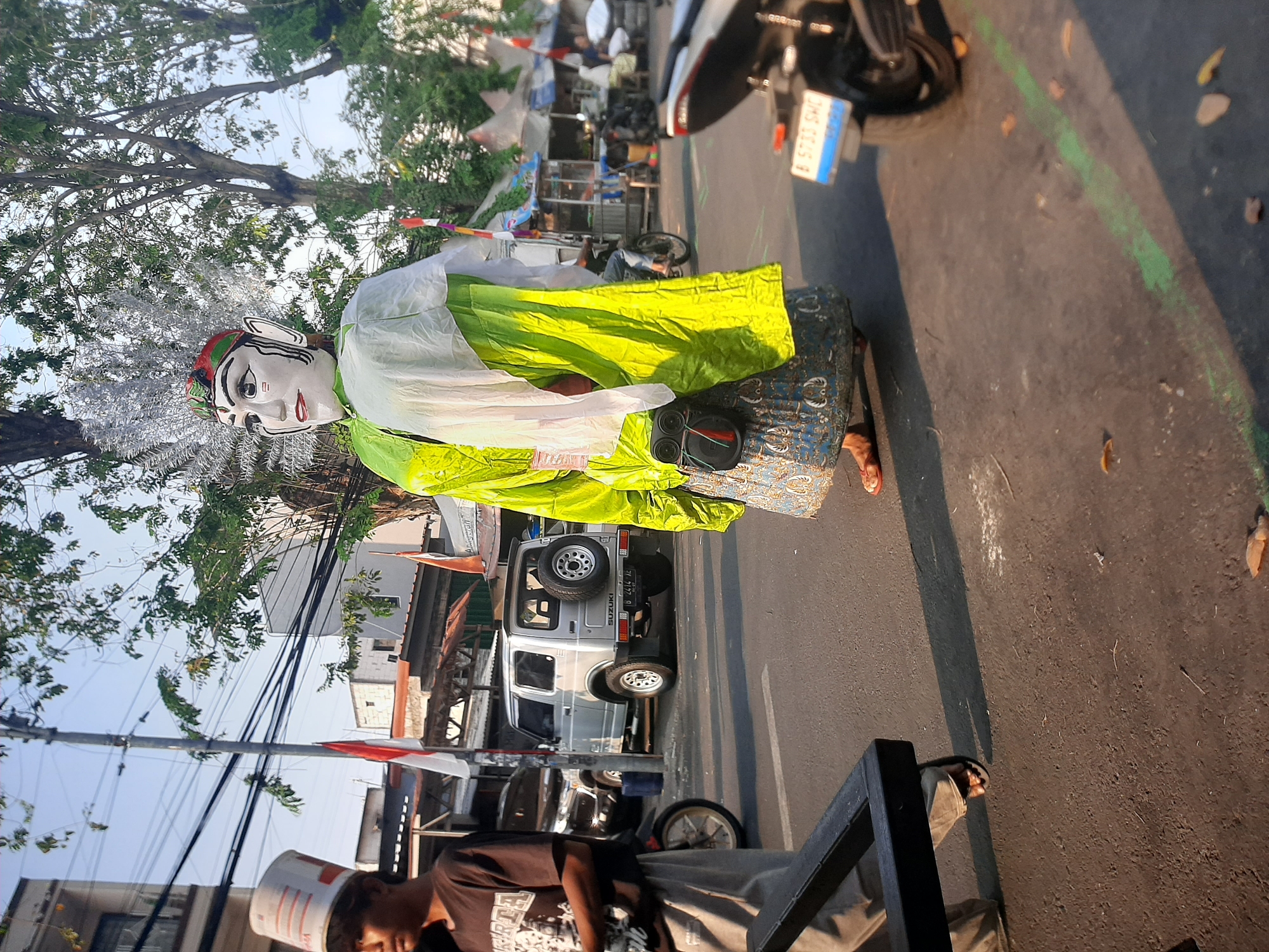 The image depicts a street scene in Central Jakarta, Indonesia, on a sunny day. In the foreground, a person wearing a brown t-shirt and light-colored pants stands on the left, partially obscured by a dark object that appears to be part of a frame or barrier. They are wearing a white bucket hat with red and orange stripes. To the right of this person is a large, traditional Indonesian "Ondel-ondel" puppet, with a white head featuring painted facial features, red and green headdress, and a flamboyant lime-green and white outfit. The puppet is adorned with a black bag on its waist. The street is paved with asphalt, and there are markings on the road, including a green line. In the background, several vehicles are visible: a silver Suzuki Jimny SUV with a spare tire on the back, a dark-colored SUV, and a motorcycle parked on the right side of the frame. Various buildings and stalls are present along the street, with trees providing shade. Indonesian flags are also visible, indicating a potential celebration or event. The overall atmosphere suggests a lively street setting with cultural elements.