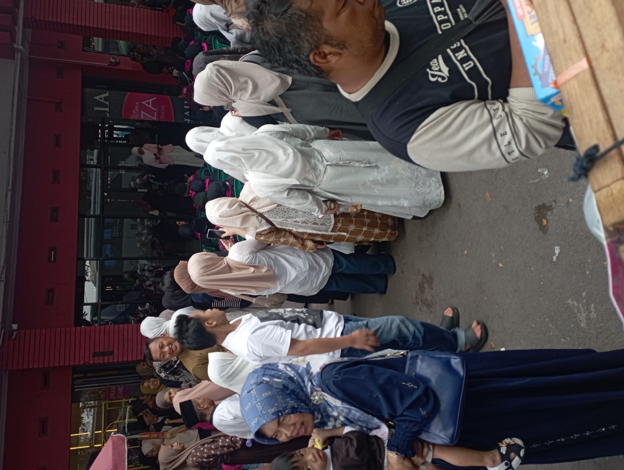The image depicts a crowded street scene in Tasikmalaya, Indonesia, likely in front of a commercial establishment. A red building with large windows and signage reading "ZA TRAVEL" is visible in the background. Numerous individuals, predominantly women wearing hijabs and modest clothing, are gathered, some appearing to be in conversation or observing something. A woman in a blue hijab is holding a young child. Men are also present, including one in the foreground wearing a black and beige t-shirt. The ground is paved and appears to be a street or pedestrian walkway. The atmosphere suggests a public gathering or a busy shopping area.