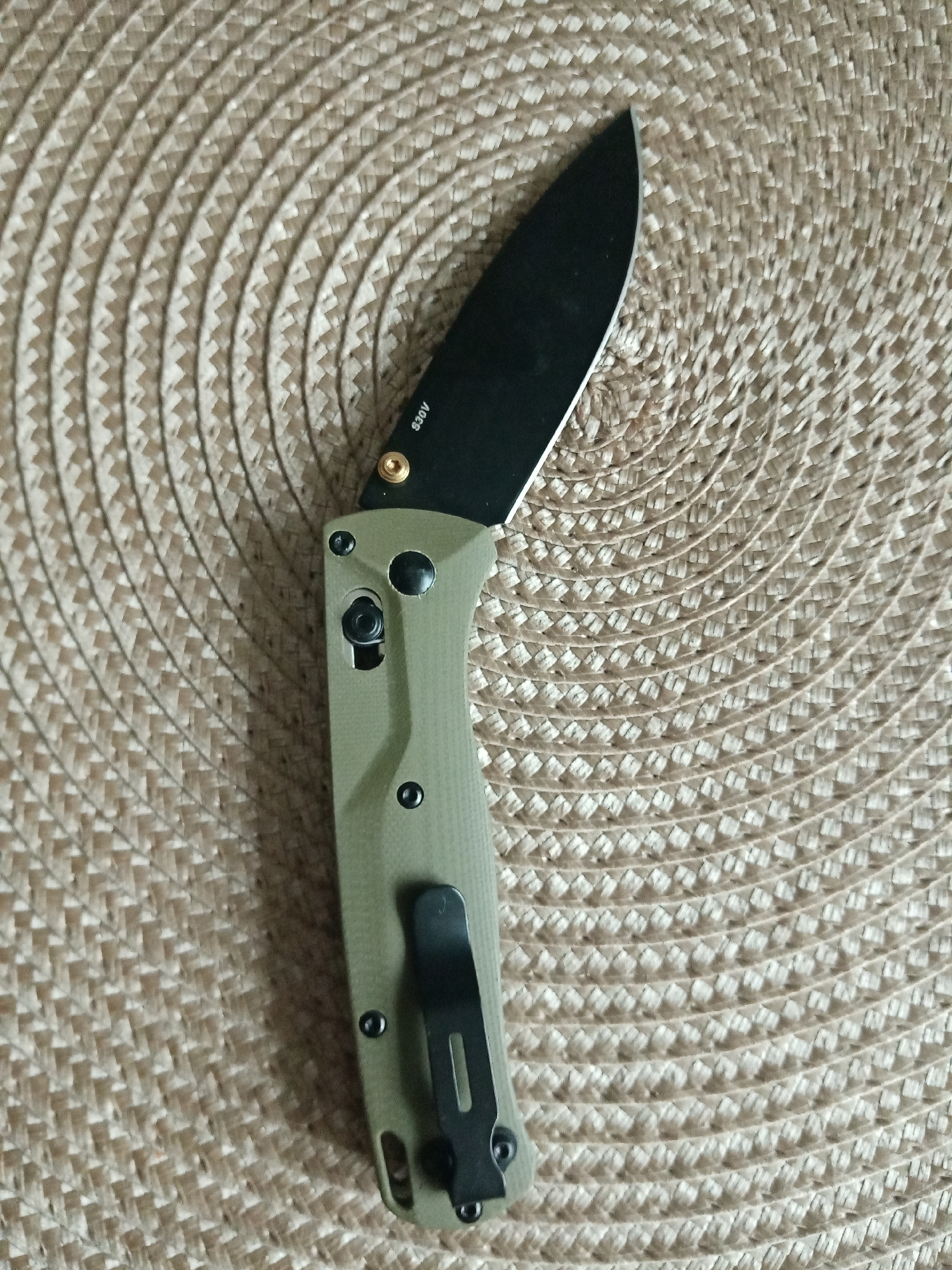 A folding knife with a black blade and an olive green handle is lying on a woven placemat. The placemat has a repeating circular pattern. "S30V" is printed on the blade near the handle. A black clip is attached to the handle.
There is no indication of this image being taken in Elektrostal, Russia, or any information about people or interactions in the image. The environment is simply the surface provided by the placemat.
