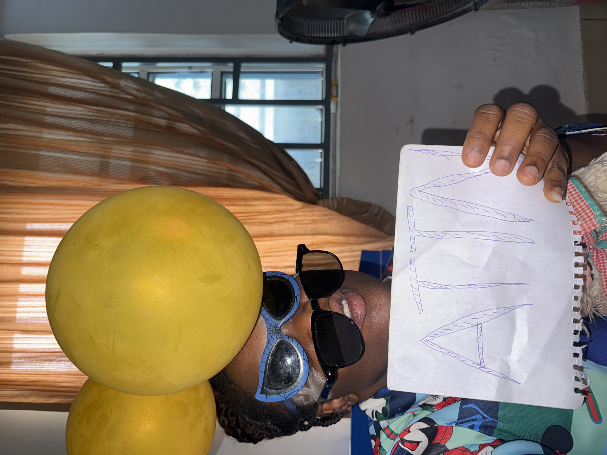 A man is shown in an indoor environment. The photograph is rotated counterclockwise. He is holding a notebook with the letters "MTN" written on it in blue ink. He is wearing sunglasses, one pair blue and the other black, and is partly obscured by two stacked yellow balloons. The background includes a window with blinds, a ceiling fan, and a patterned blanket. The location is identified as Sabon Tasha, Nigeria.