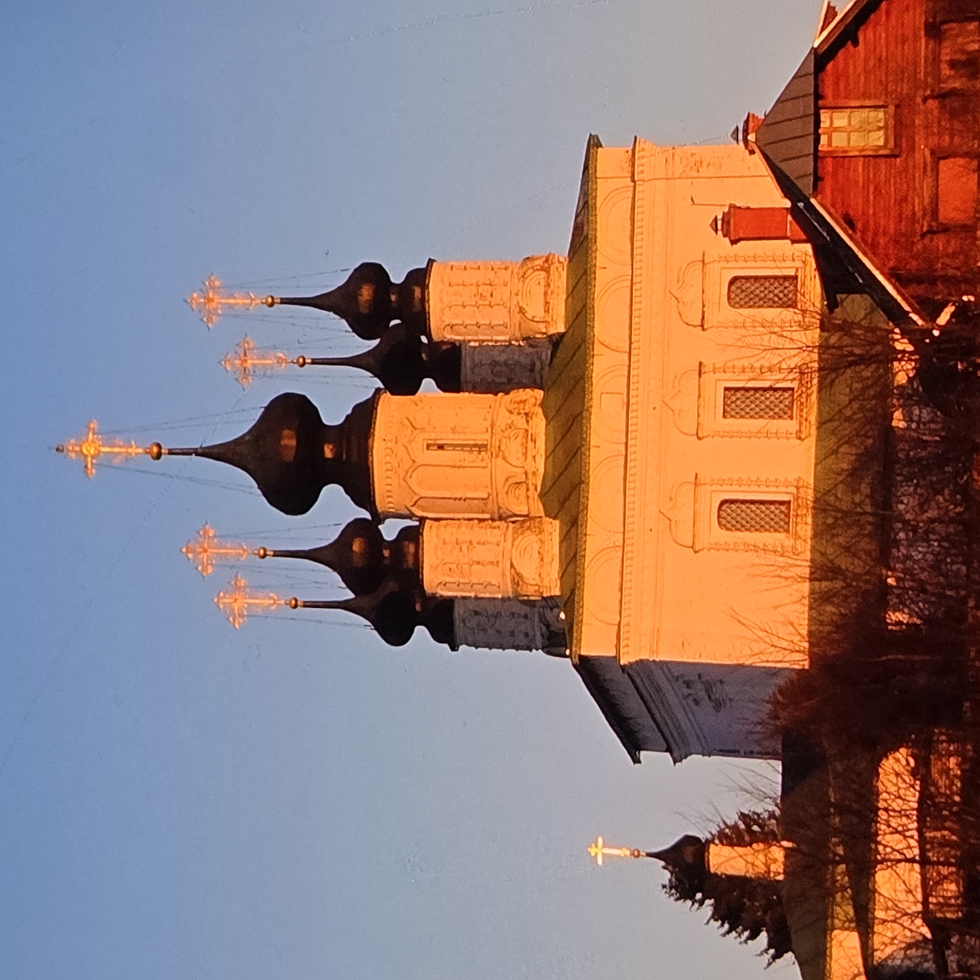 The image depicts the golden-domed Transfiguration Cathedral in the city of Vladimir, Russia, during a sunset. The cathedral's white brick facade is bathed in the warm, orange glow of the setting sun, highlighting its architectural details, including arched windows and decorative carvings. Several onion domes, topped with ornate crosses, rise from the structure. To the left, a smaller bell tower with a single cross is visible, also illuminated by the sunset. In the foreground, a red wooden building with a dark roof and a window is situated, with its side facing the viewer. The scene is framed by a clear, pale blue sky, with silhouetted bare tree branches in the lower part of the image, casting shadows. The overall impression is one of serene beauty, with the light of the sunset accentuating the spiritual and historical significance of the cathedral.