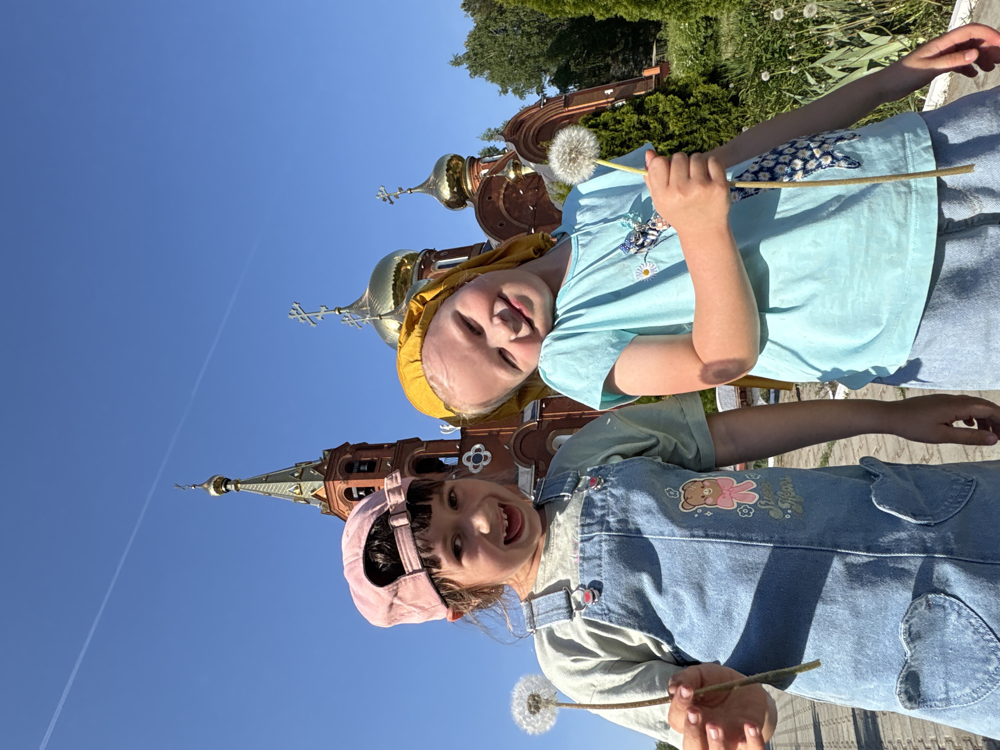 Two young girls are holding dandelions in front of a Russian Orthodox church in Chaikovsky, Russia. The girl on the left is wearing a pink cap and a denim outfit with a cartoon bear design. The girl on the right is wearing a yellow headscarf and a light blue shirt. The church has golden domes and red brick walls and stands against a clear blue sky.