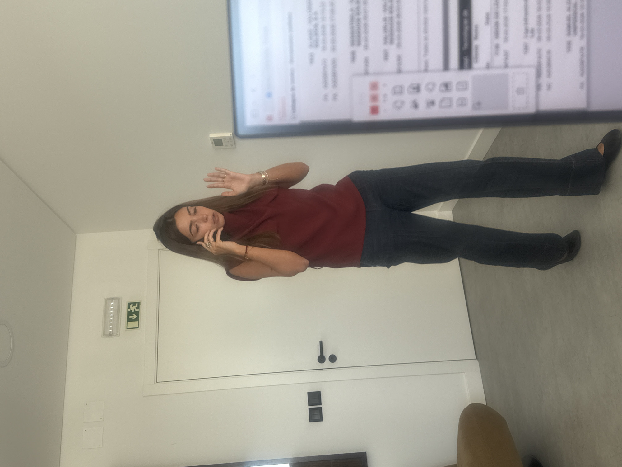 The image depicts an indoor office or commercial space located in Torres Vedras, Portugal. A single adult female stands centrally, facing slightly towards the right side of the frame. She has long, straight brown hair and is dressed in a sleeveless maroon top with a high collar, dark blue trousers, and dark flat shoes. Her left hand holds a dark-colored smartphone to her left ear, and her mouth is open, indicative of speaking. Her right hand is raised to chest height, palm facing forward, with fingers slightly apart, in a gestural motion. Multiple bracelets are visible on her right wrist, and a ring is present on her left hand.
The background features a white wall with a white door, equipped with a black horizontal handle and a circular knob positioned below it. To the left of the door, two black square electrical wall fittings are mounted. Above the door, a rectangular LED light fixture is installed next to a green emergency exit sign, which displays a running figure and an arrow pointing diagonally downward and to the right. Further right on the wall, a small white wall-mounted control panel, possibly a thermostat, is visible. The floor is a uniform light grey.
On the far left, a section of a brown upholstered armchair with a dark base is partially visible. On the right foreground, a computer monitor partially obstructs the scene, displaying a software interface with multiple lines of text, numbers, and icons on a light background.